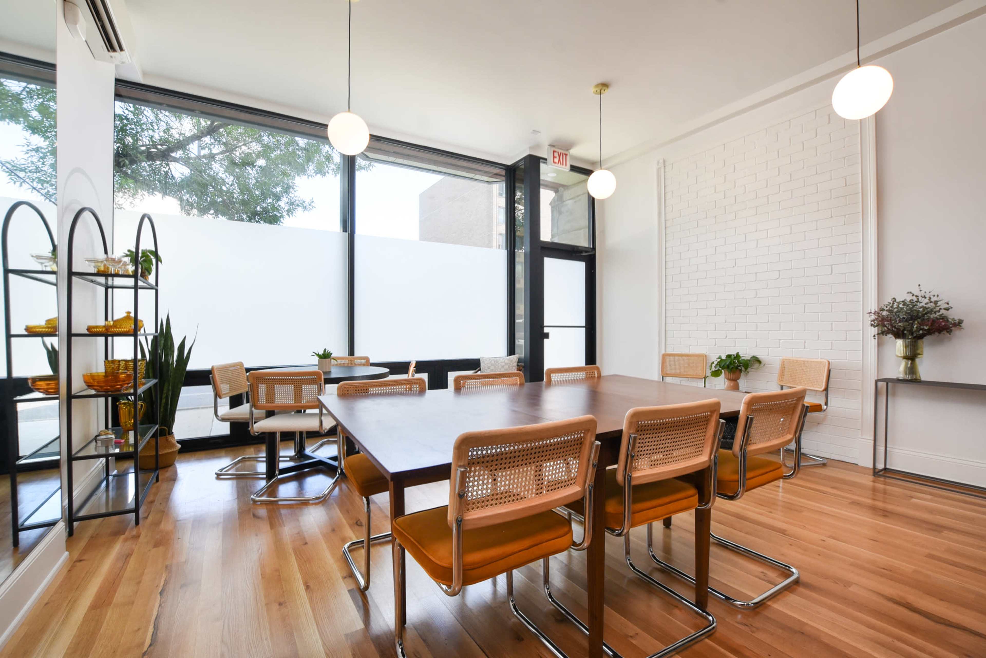 The image shows a modern dining area with a large wooden table surrounded by eight chairs, large windows letting in natural light, and a minimalist decor featuring plants and shelves.
