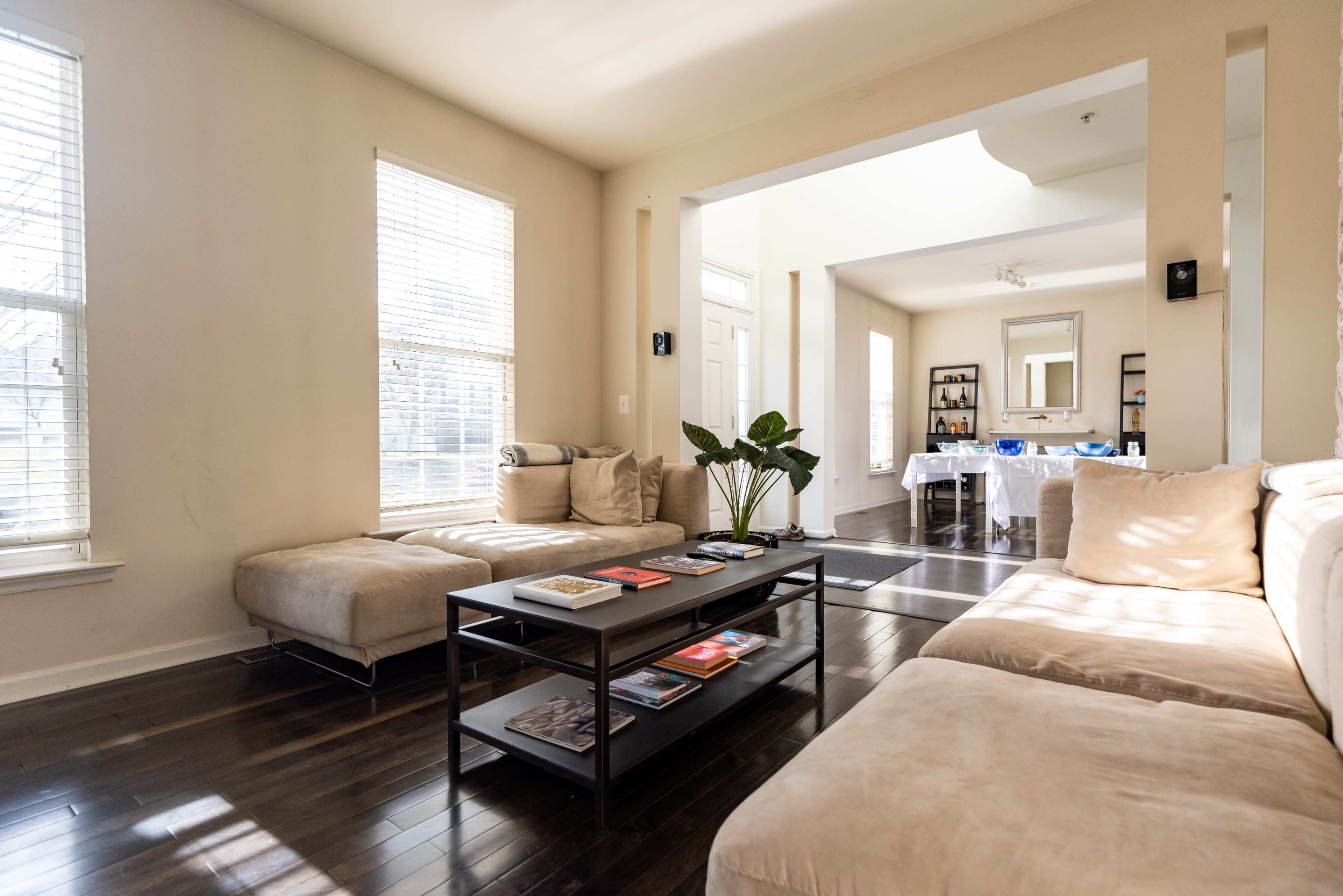 A bright living room with two sofas, a coffee table stacked with magazines, and large windows letting in natural light.
