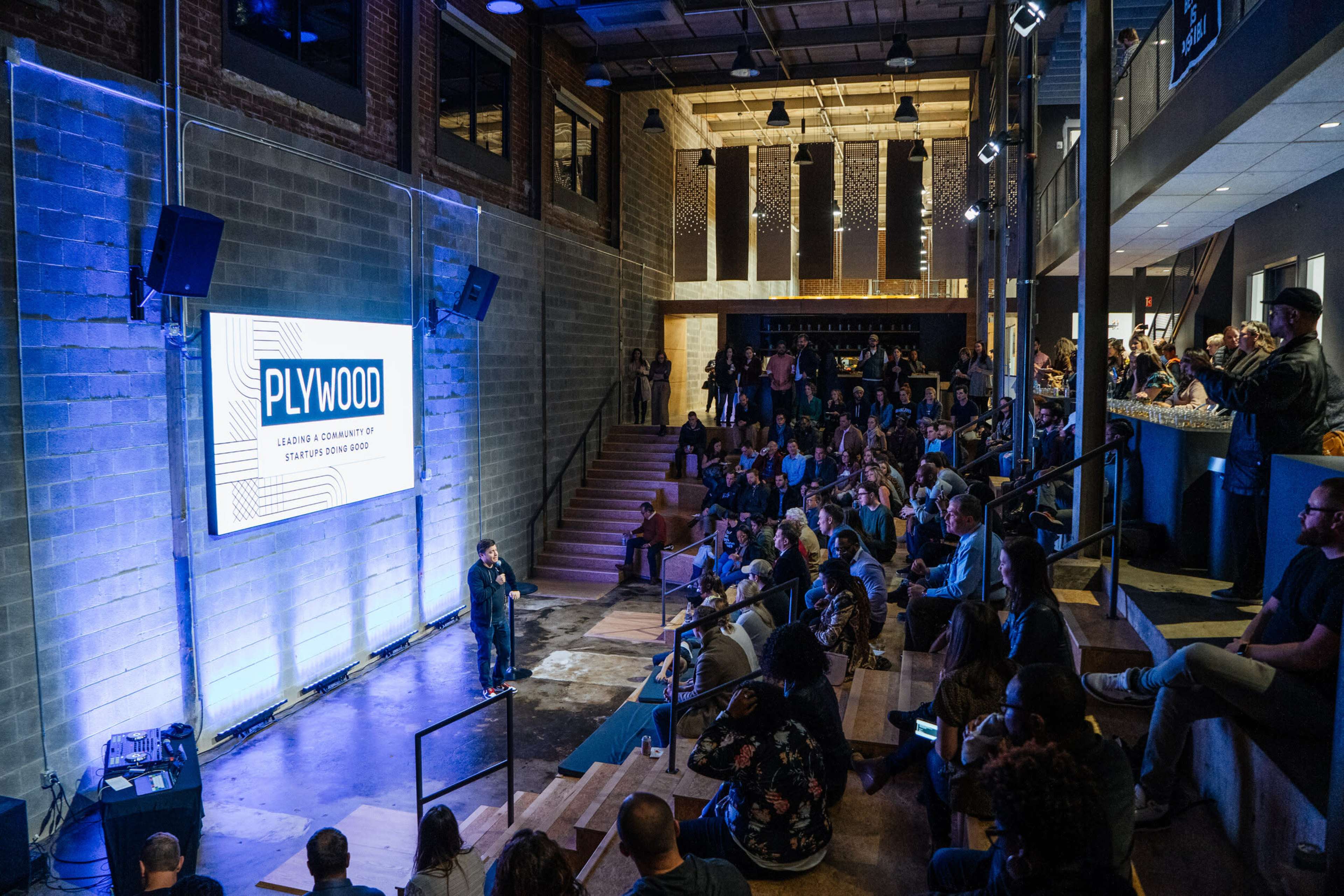 A speaker stands on a stage in front of a large audience seated in a modern venue with industrial decor, while a screen displays the word "PLYWOOD."