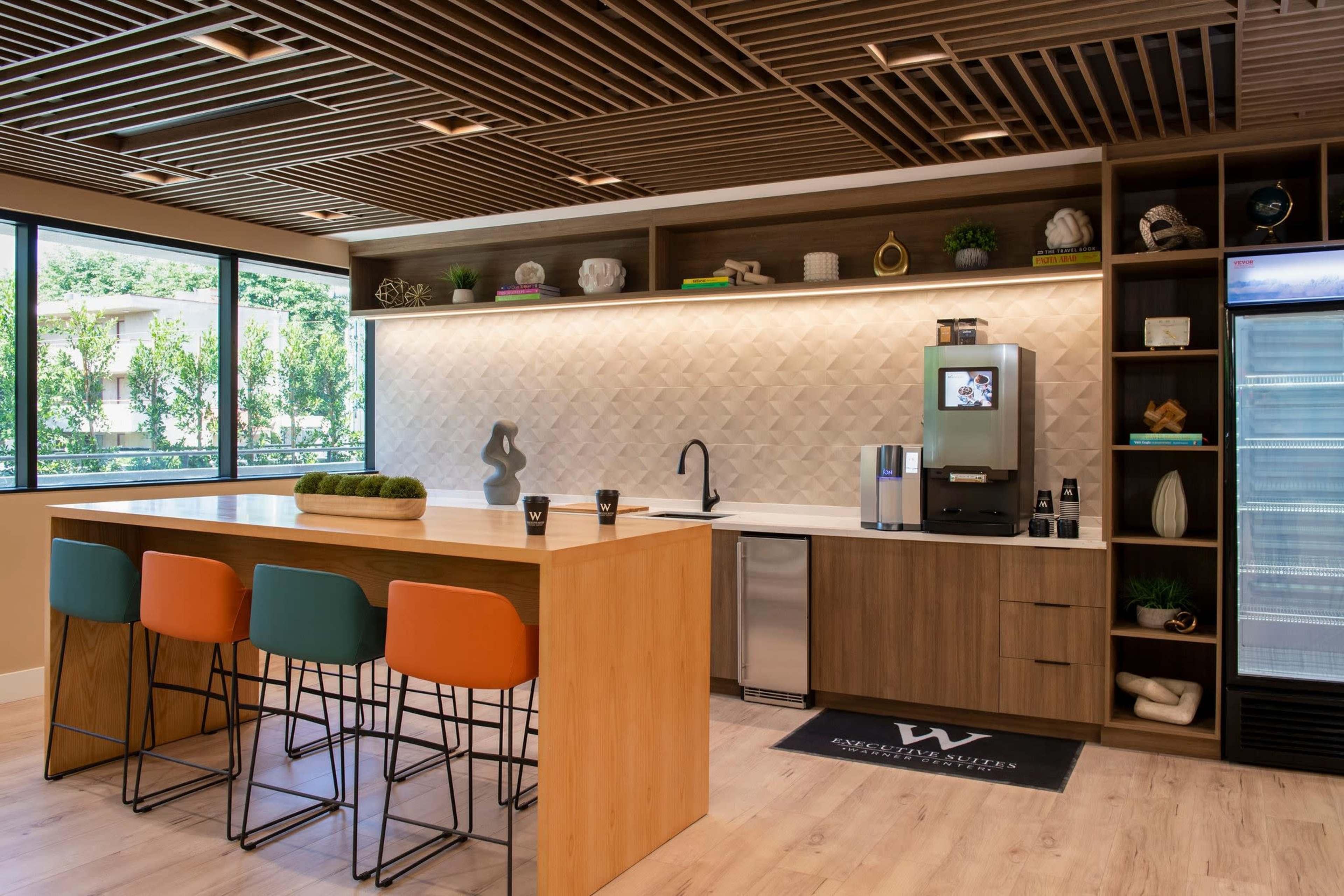 The image shows a modern kitchen area featuring a wooden island with colorful bar stools, a sleek countertop, and an organized shelf with decorative items and a coffee machine.