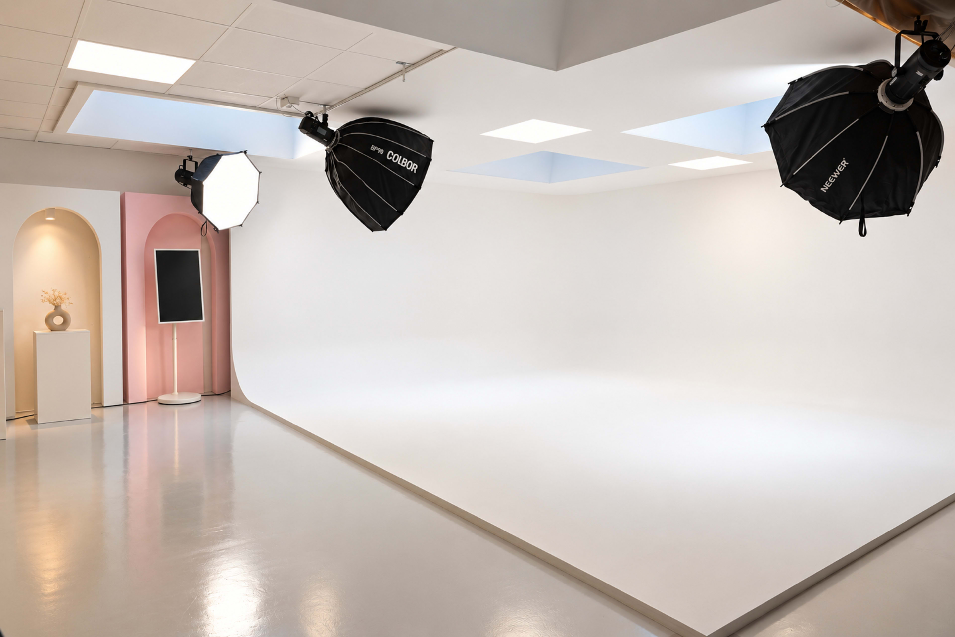 The image shows a well-lit photography studio featuring a seamless white backdrop, two large softbox lights, and a minimalist decorative element.