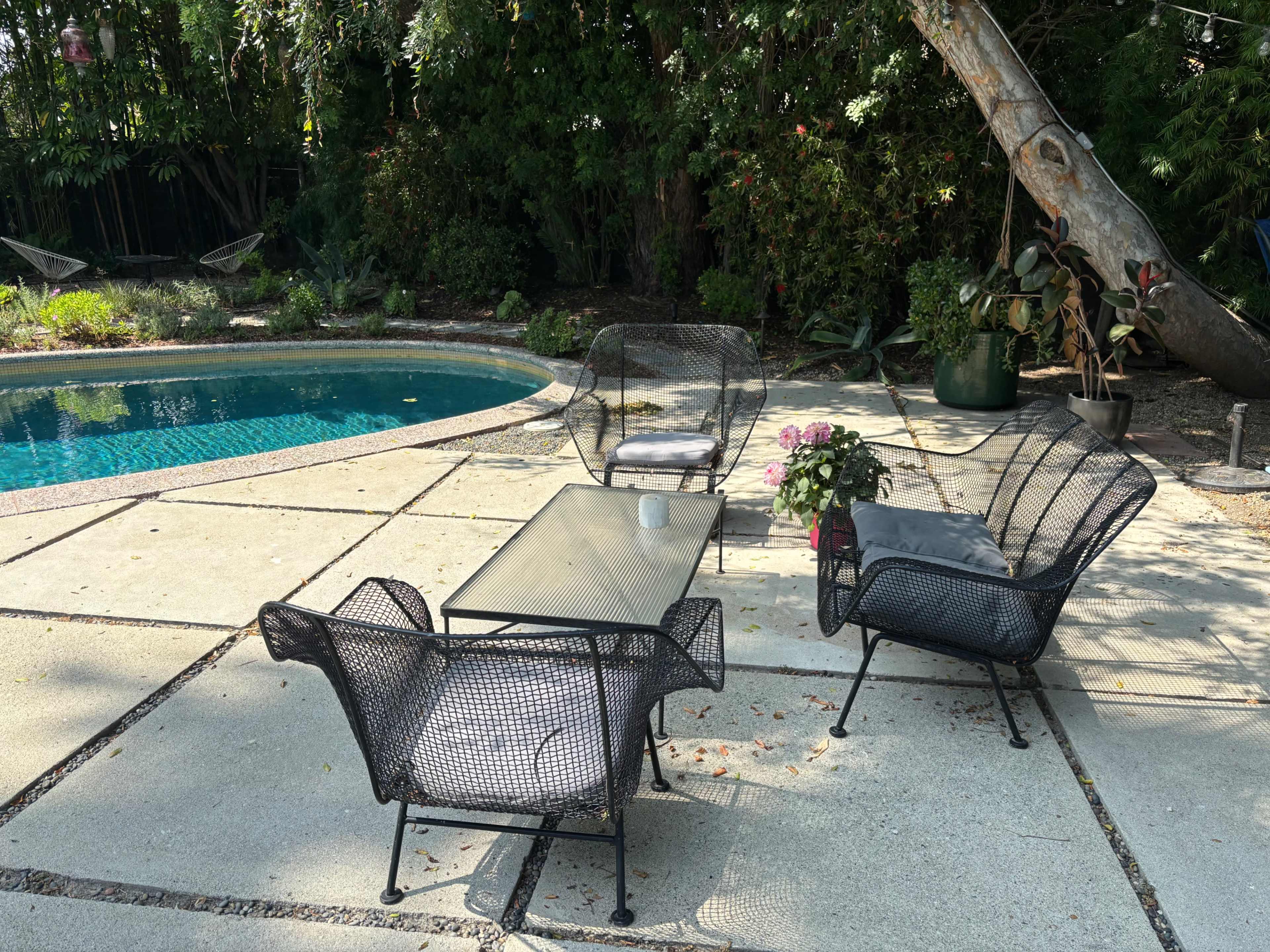 A small outdoor seating area features four black wire chairs around a glass-topped table beside a blue swimming pool, surrounded by lush greenery.