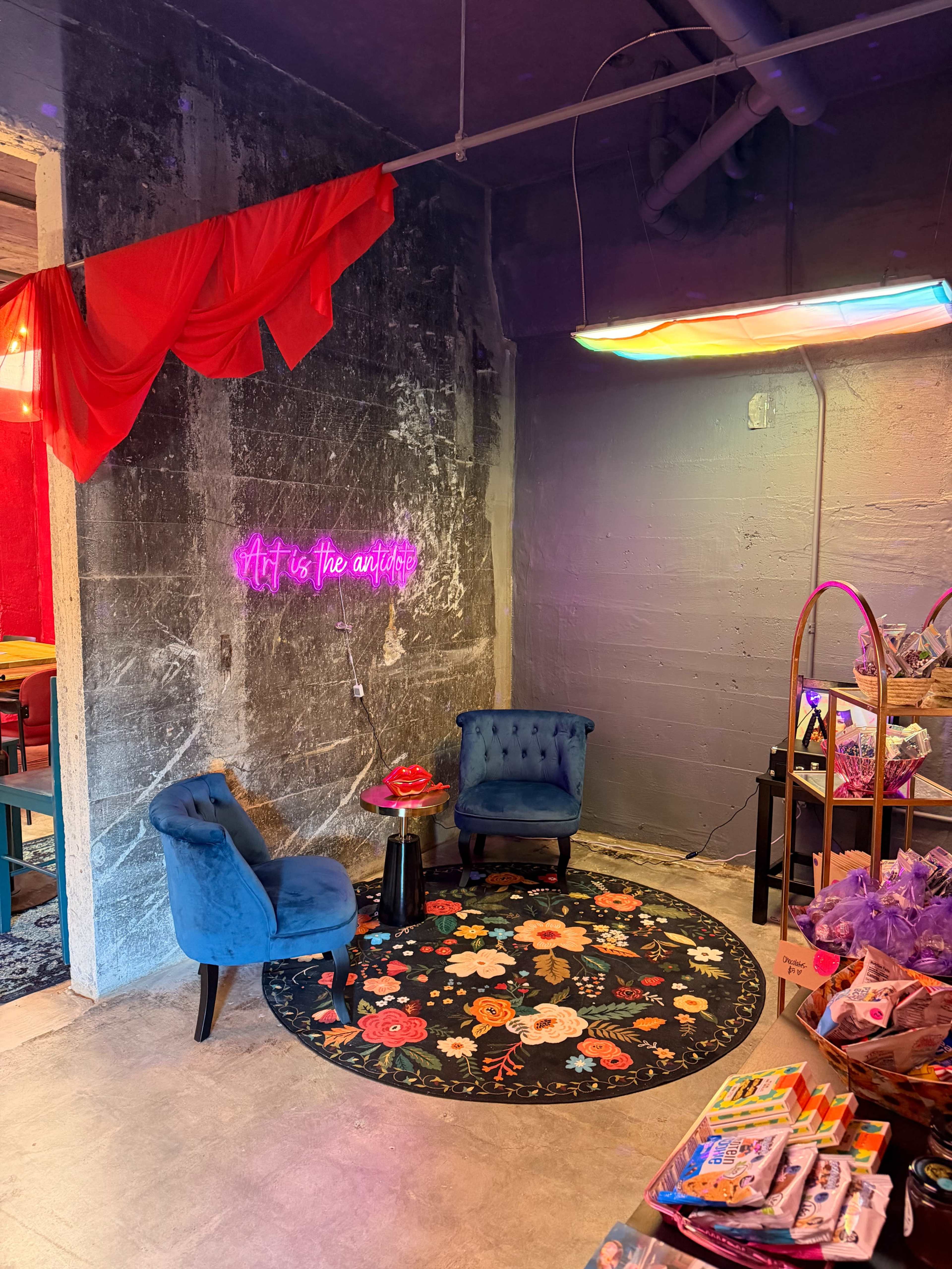 The image depicts a small seating area with two blue chairs, a circular floral rug, and a neon sign that reads "Art is the antidote," alongside shelves filled with colorful products.
