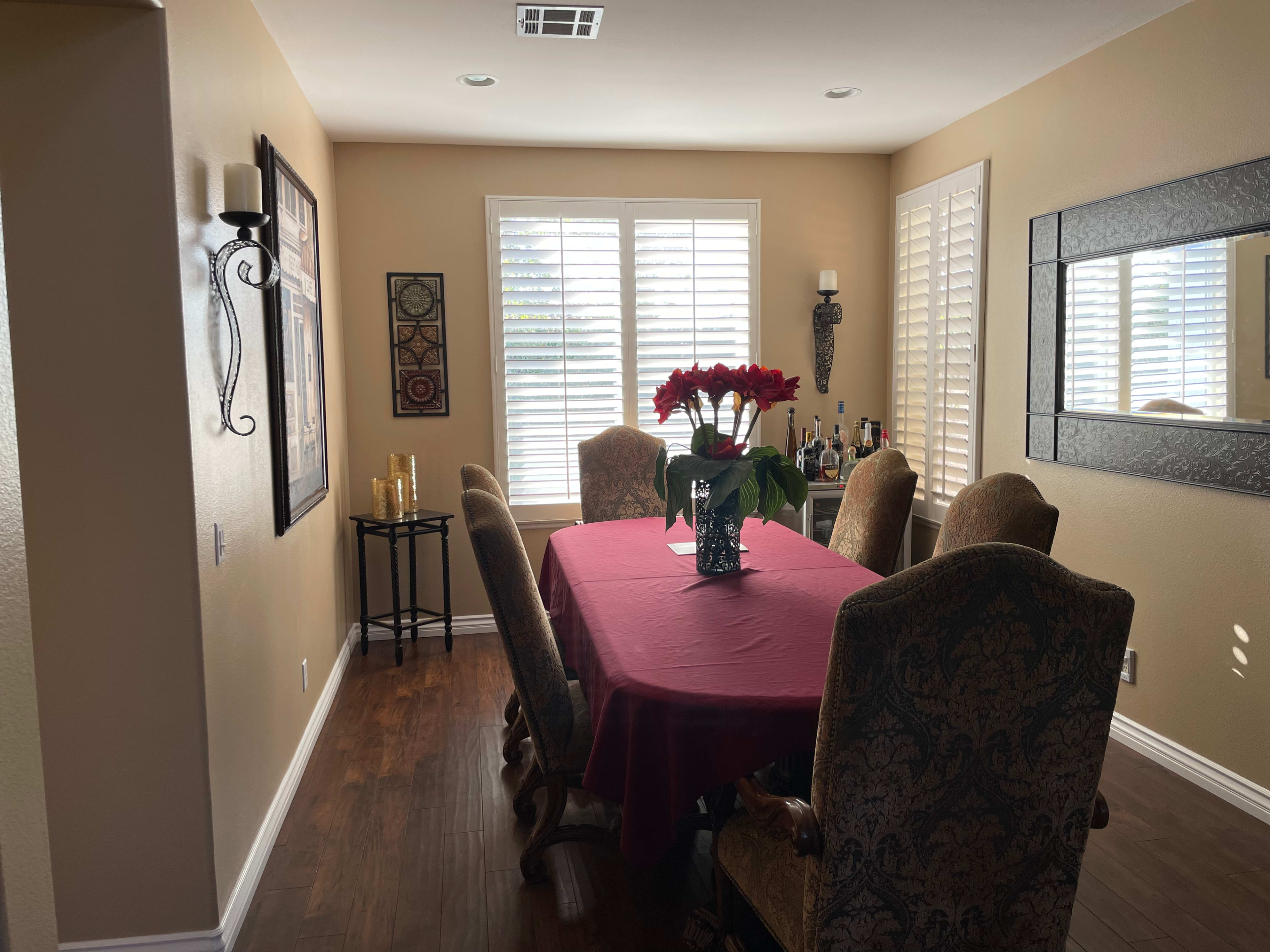 A dining room features a rectangular table with a red tablecloth and a vase of flowers, surrounded by upholstered chairs and illuminated by natural light through window shutters.