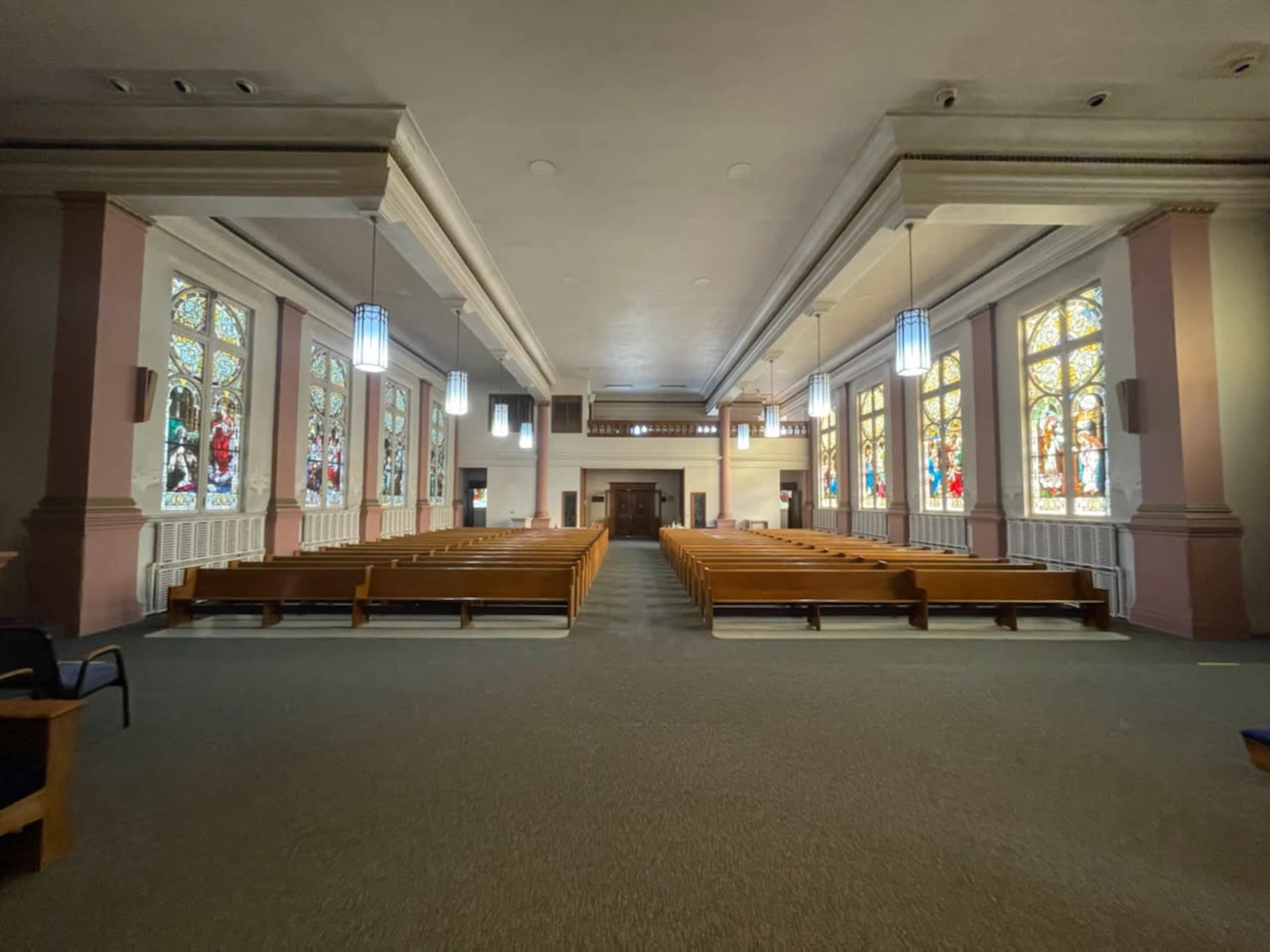 The image shows the interior of a church with wooden benches arranged facing an altar, illuminated by stained glass windows on either side.