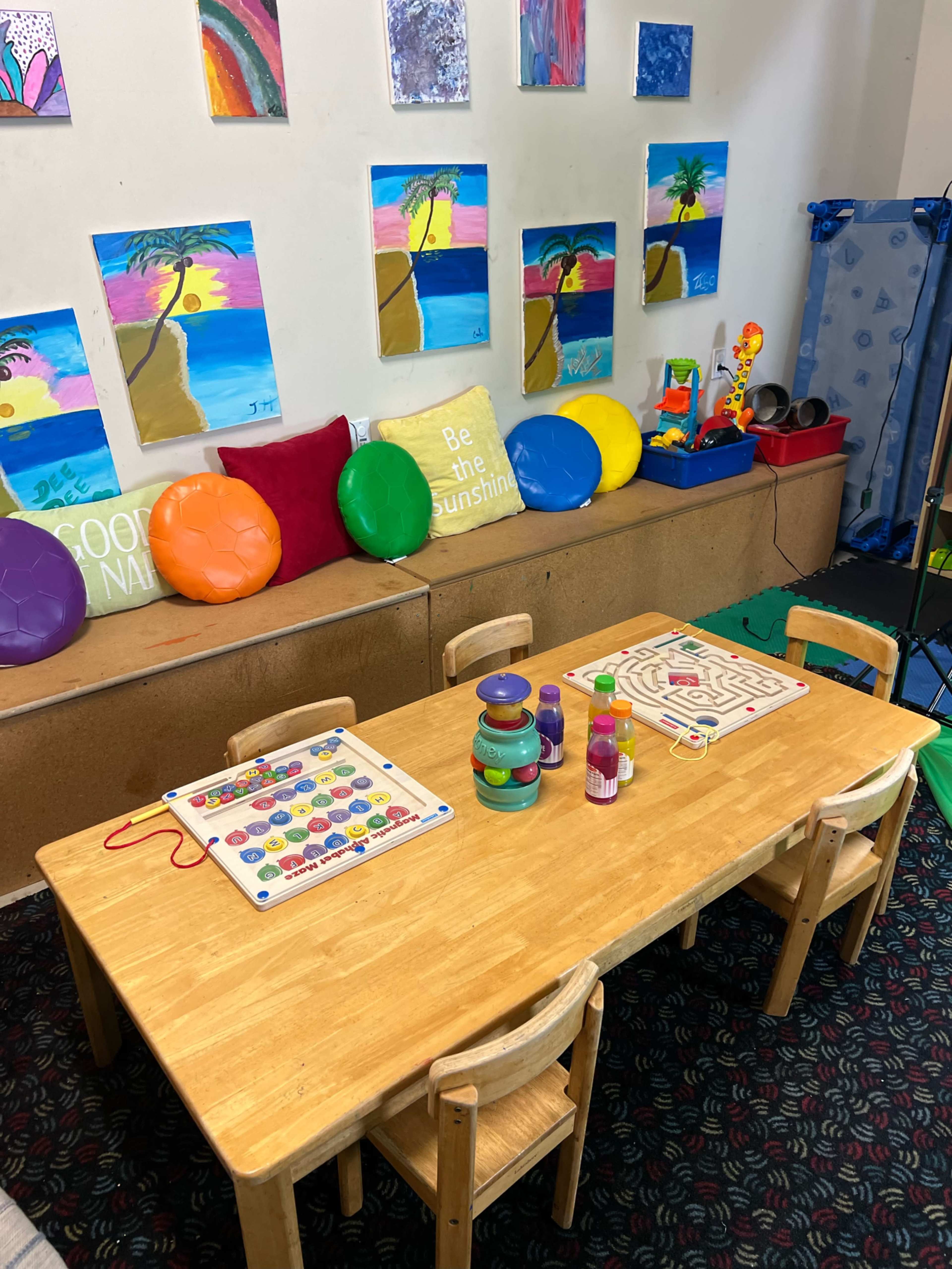 A classroom corner features a wooden table with learning games and colorful toys, surrounded by artwork depicting tropical scenes on the walls.
