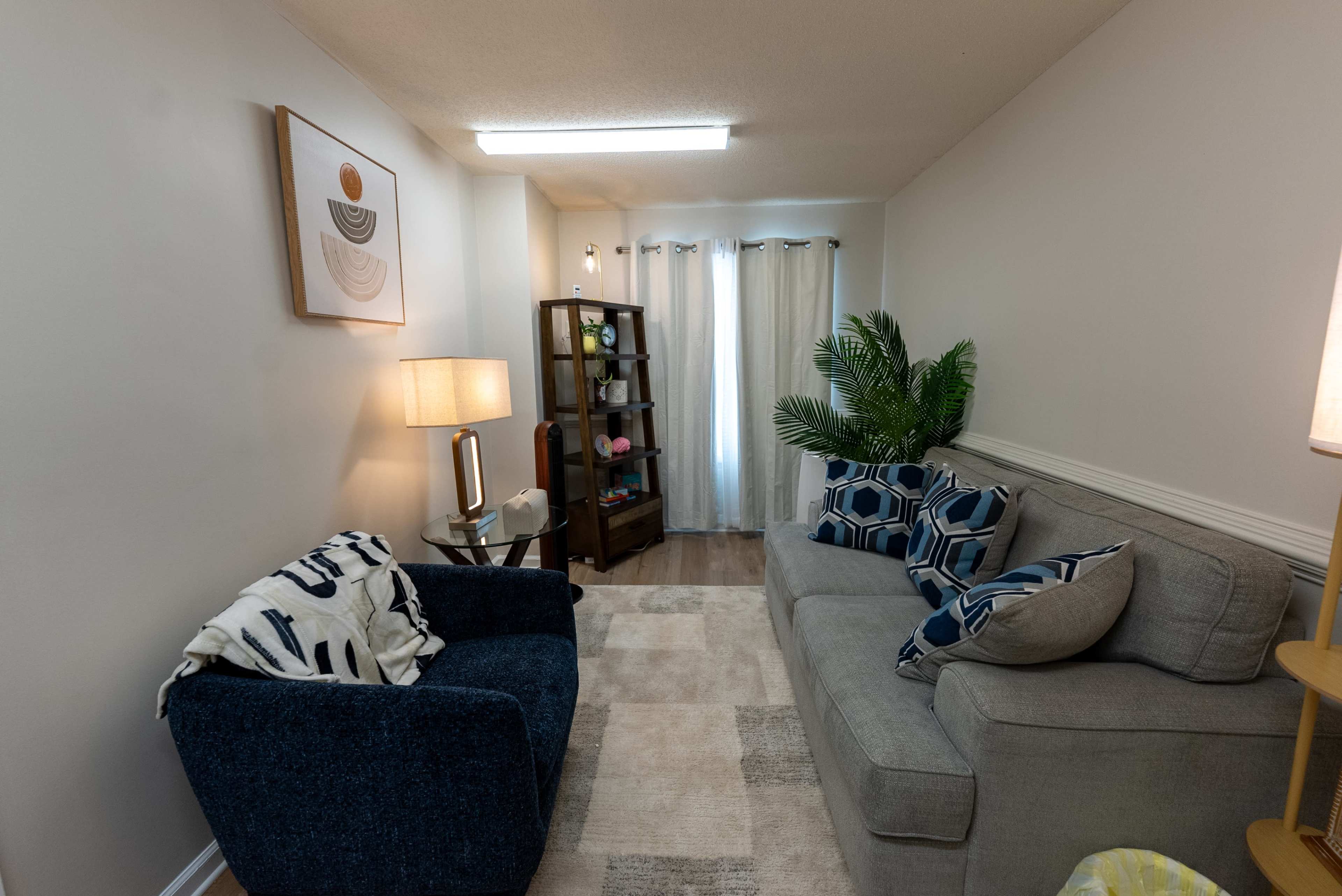 The image shows a cozy living room with a blue armchair, a gray sofa with decorative pillows, a wooden cabinet, and a lamp, illuminated by natural light from a window.