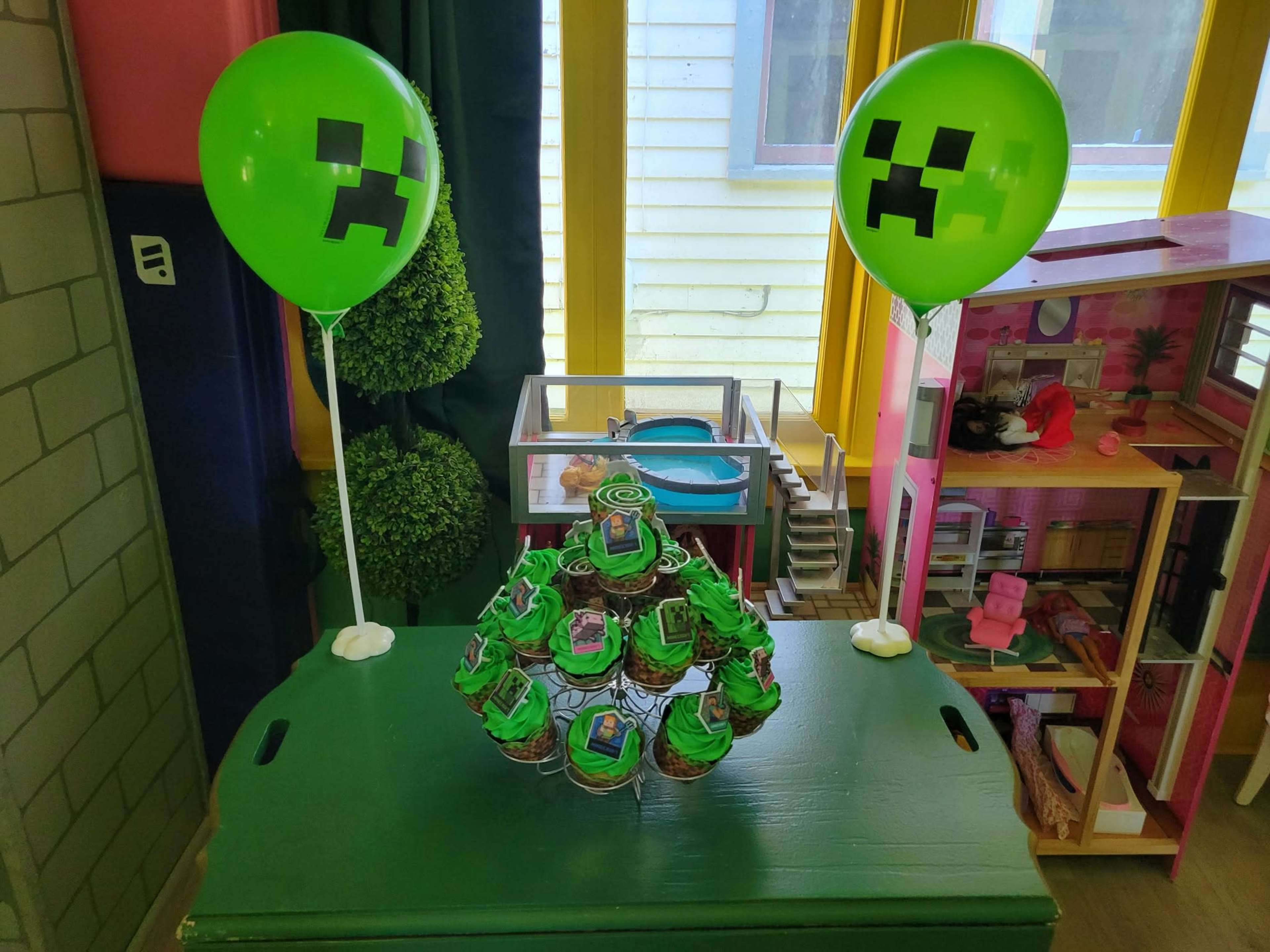 The image shows a display featuring a green Minecraft-themed cupcake tower surrounded by two green balloons with Creeper faces on them.
