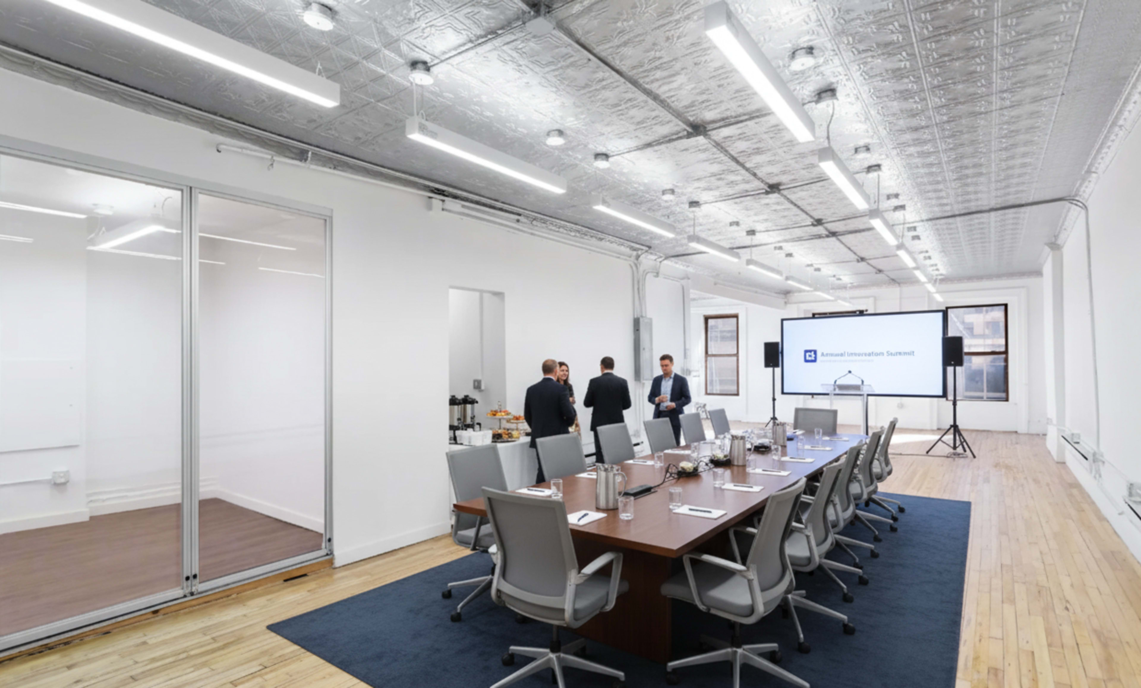 A modern conference room features a long wooden table surrounded by chairs, with a large screen and a few people engaging in conversation.