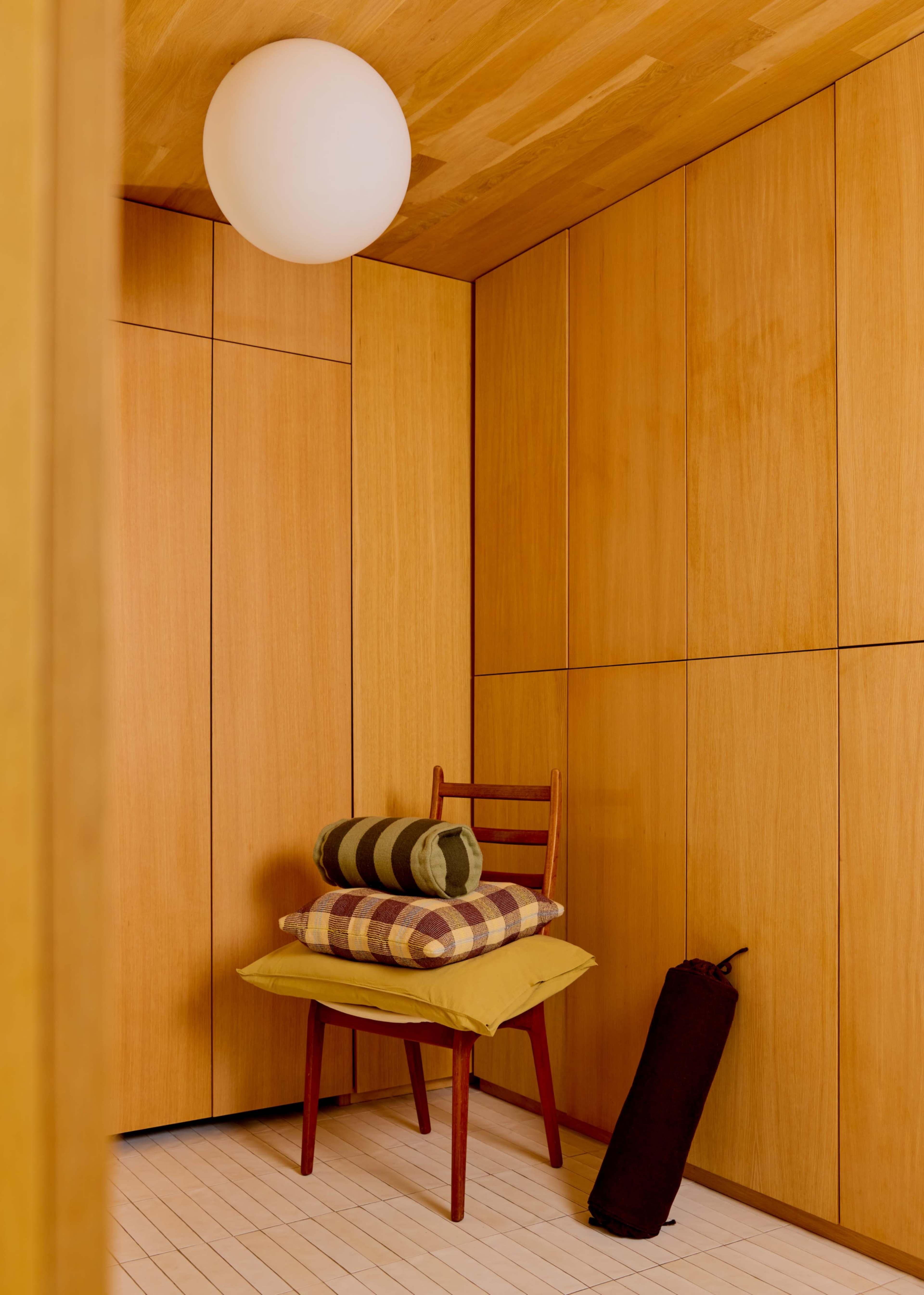 A wooden corner features a chair stacked with colorful cushions and a rolled blanket beside it, illuminated by a spherical ceiling light.