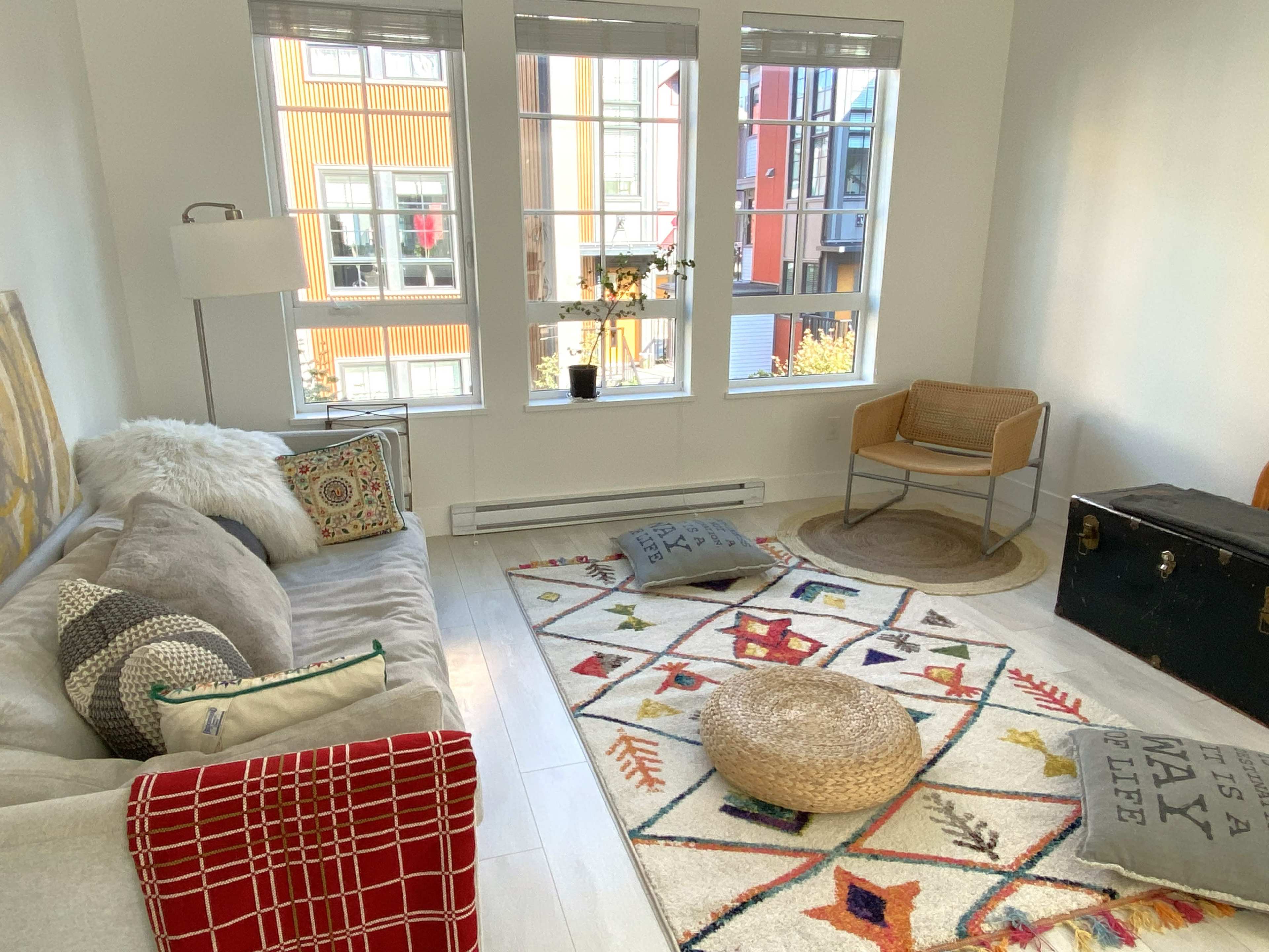 A bright living room features a light sofa, a large area rug with geometric patterns, and large windows showcasing an outside view.