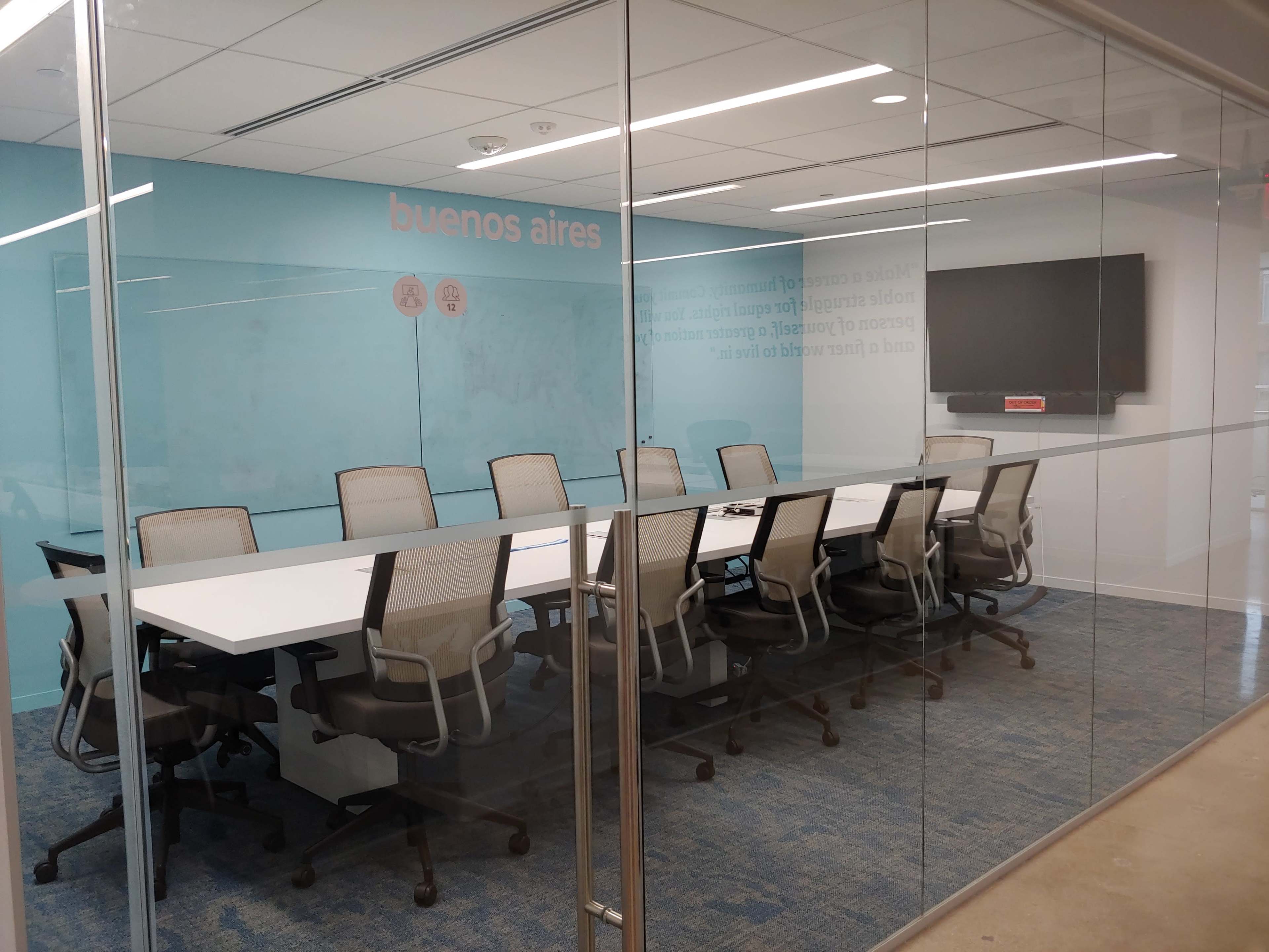A conference room with a long white table and several gray office chairs is enclosed in glass walls, featuring a TV on the wall and light blue decor.