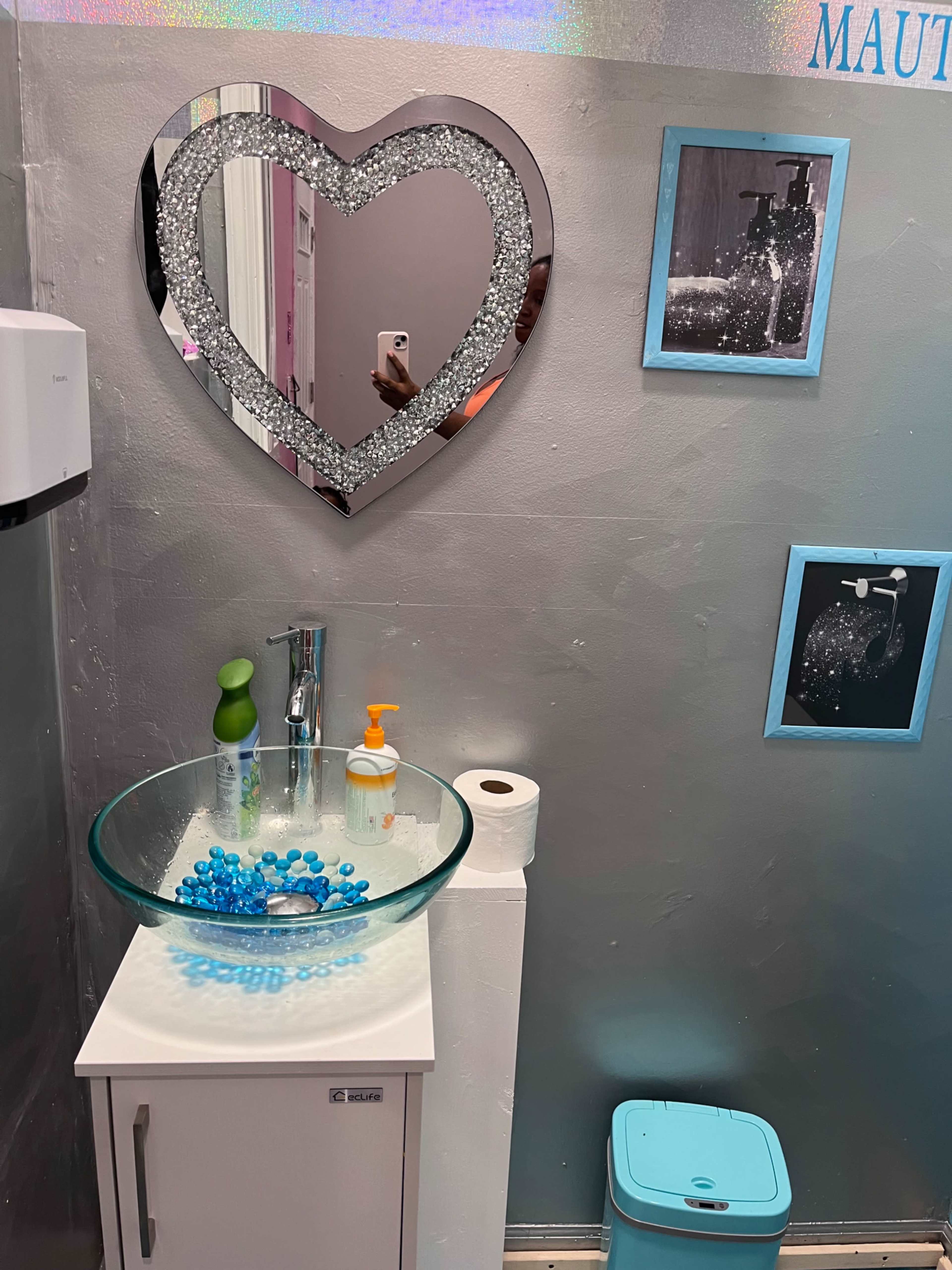 A modern bathroom with a heart-shaped mirror above a round glass sink, accompanied by a soap dispenser, a roll of toilet paper, and a small trash bin.