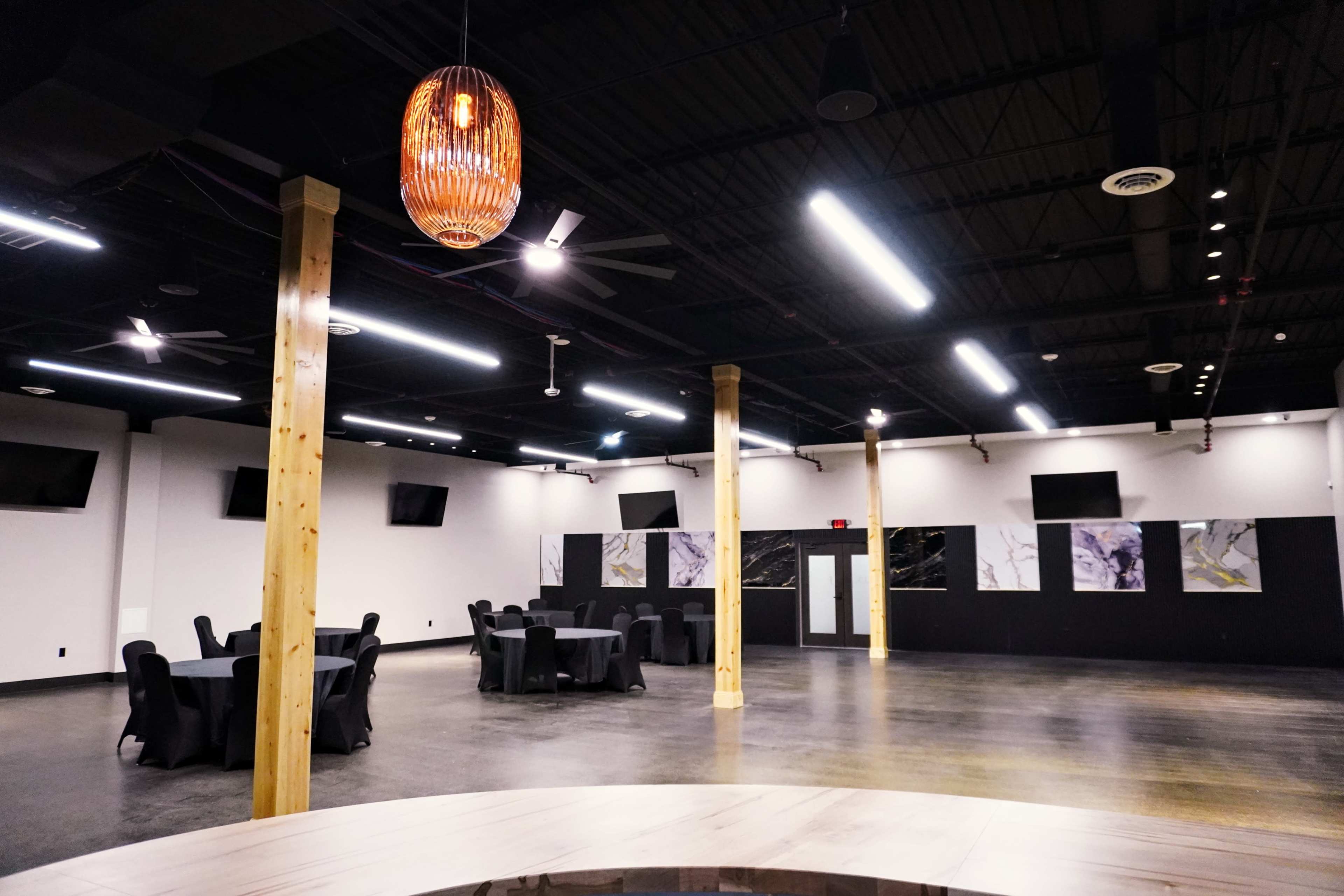 The image shows a spacious event hall with wooden pillars, black tablecloths on round tables, and abstract artwork on the walls.