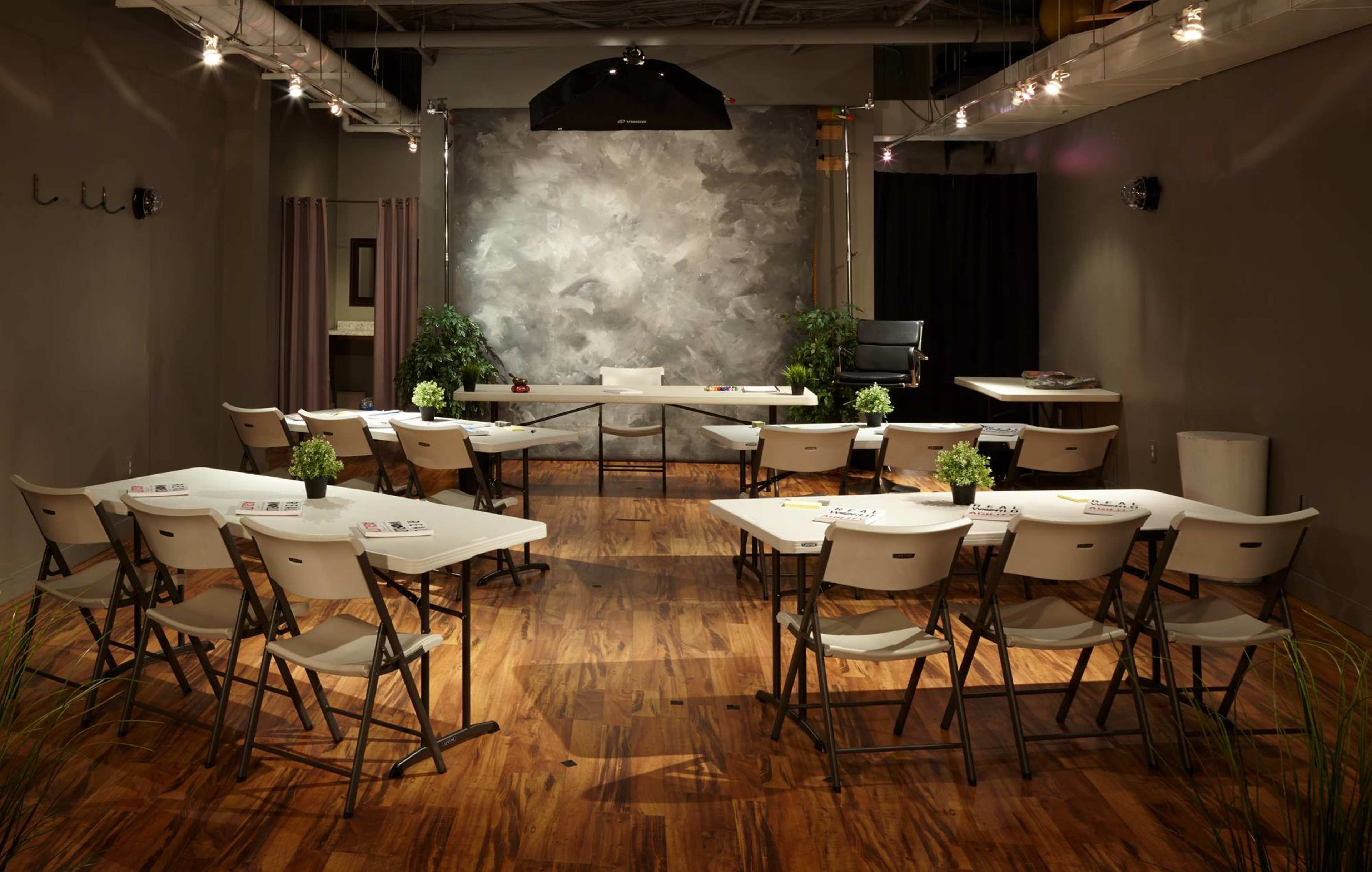 A training or meeting room is set up with rows of chairs and tables, featuring a gray backdrop and plants, illuminated by overhead lighting.