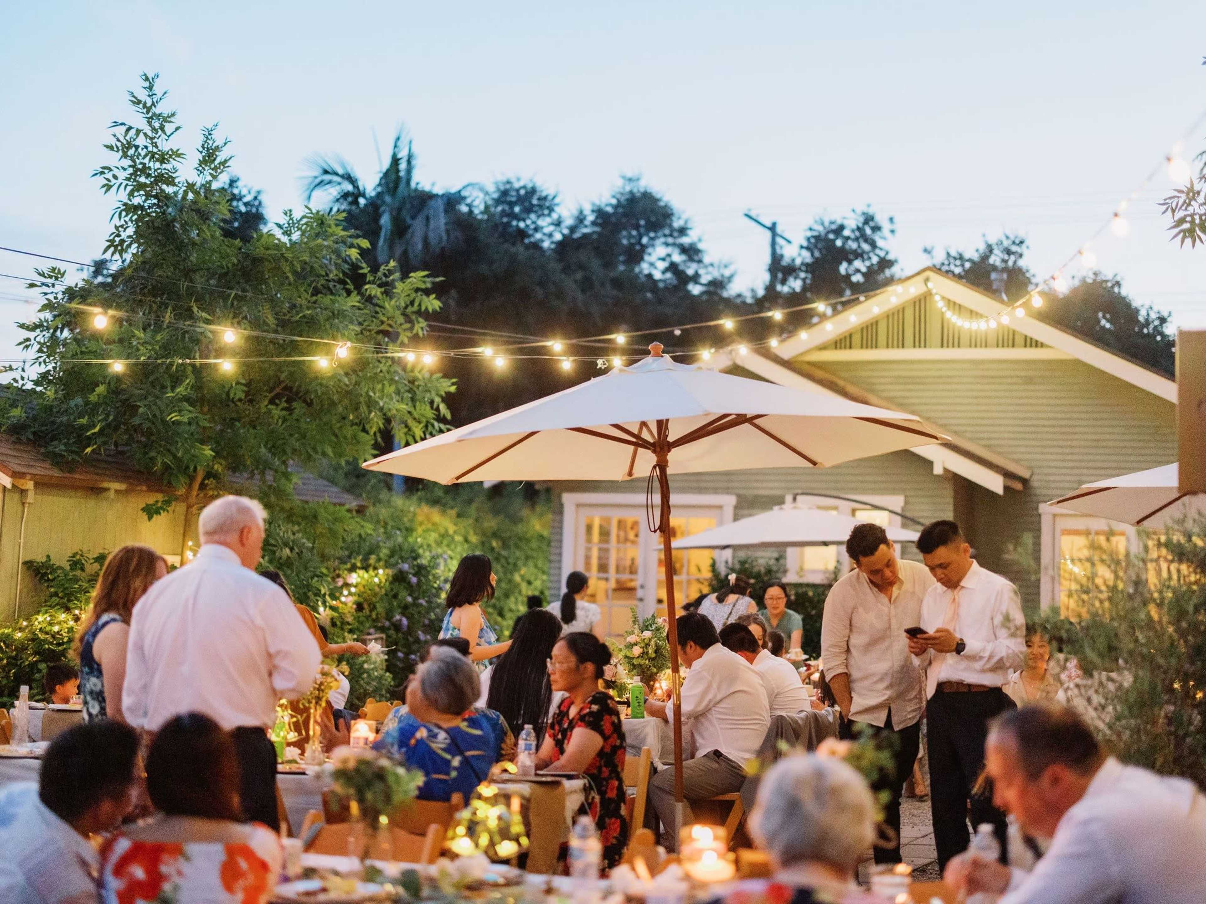 A garden dinner party is happening outdoors with guests seated at tables under string lights and umbrellas.