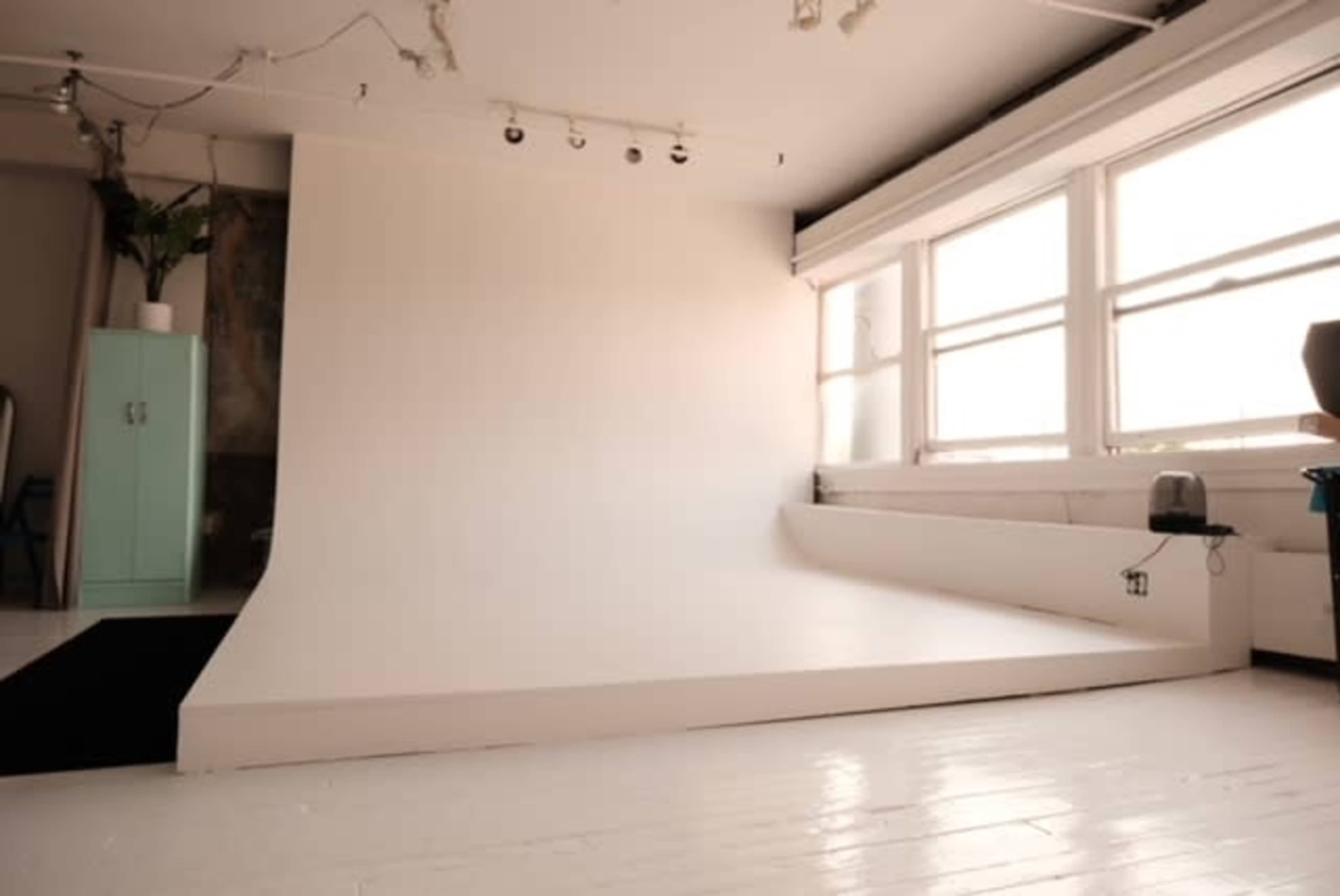 The image shows a minimalist photography studio with a white seamless backdrop, large windows letting in natural light, and wooden floors.