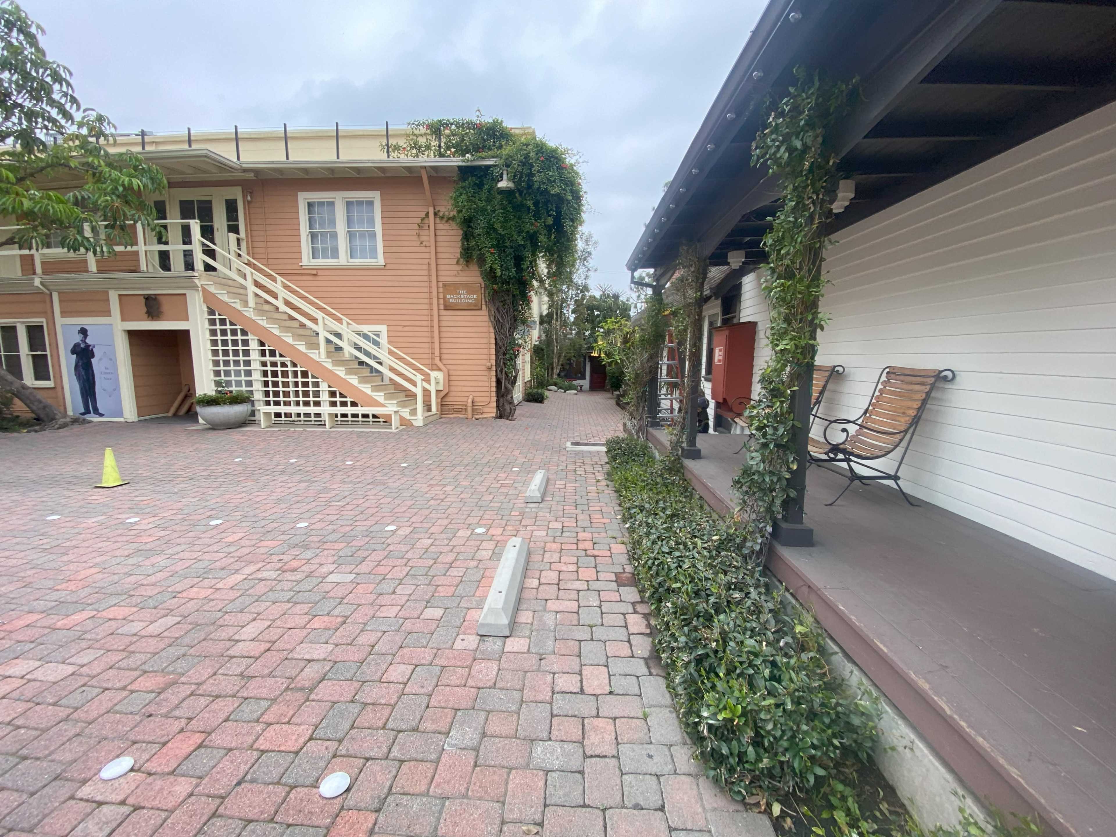 The image shows a cobblestone pathway between two buildings, one with a staircase and the other featuring a bench and greenery.