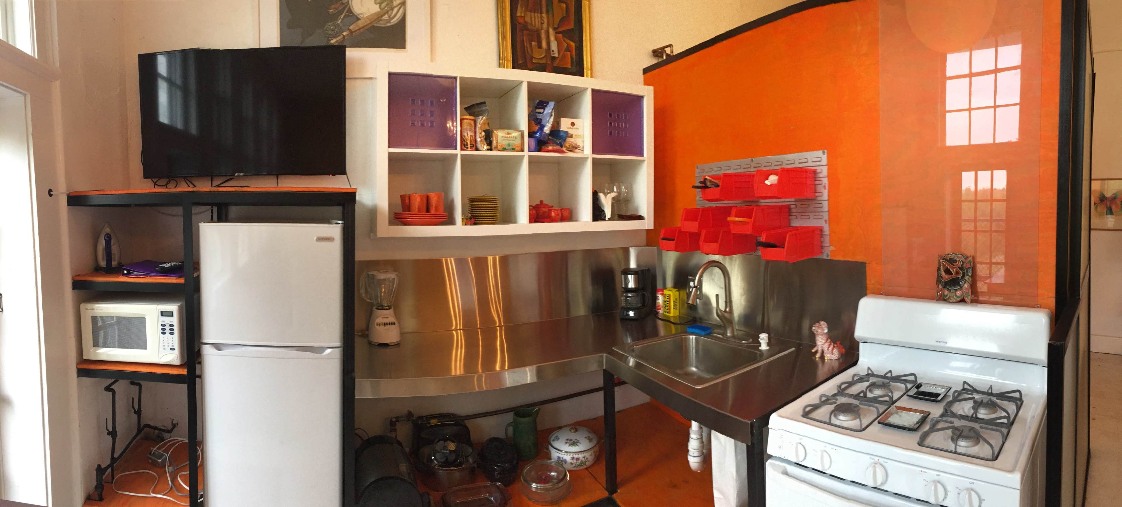 A compact kitchen with stainless steel countertops, a stove, a refrigerator, a microwave, and colorful shelving units.