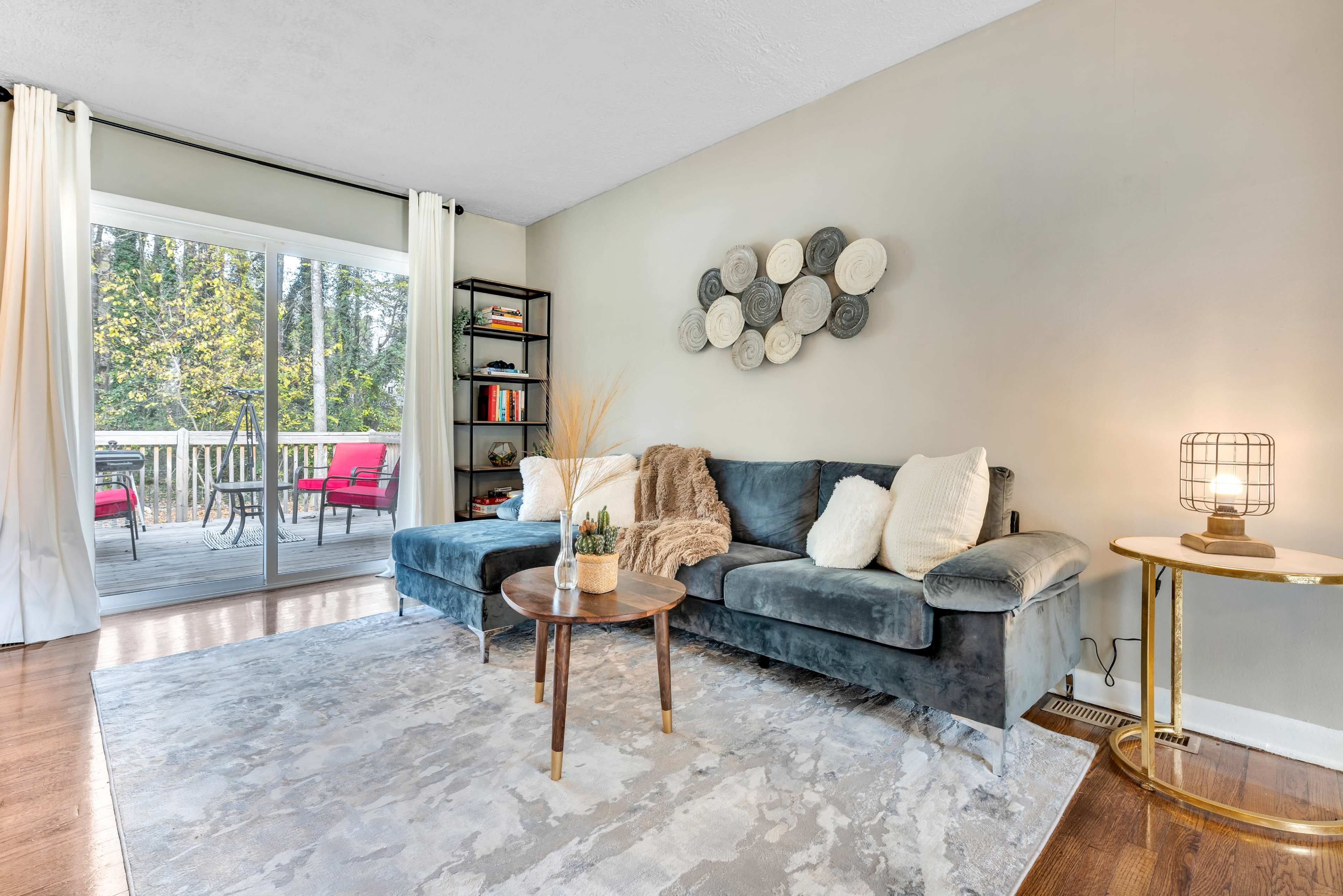 A cozy living room with a gray sectional sofa, a round coffee table, and a large window that provides a view of an outdoor deck with red chairs.
