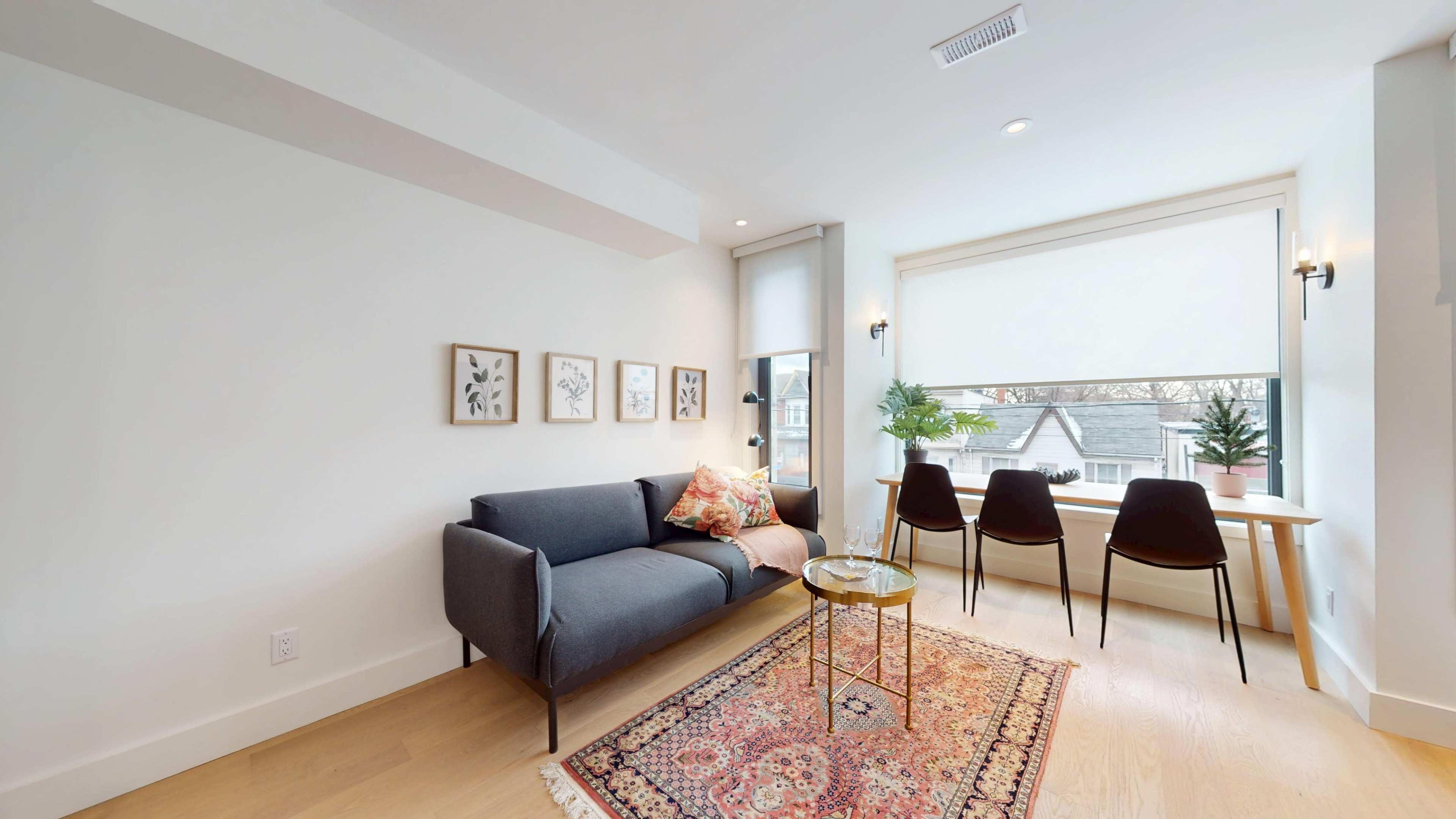 The image shows a modern living space with a gray sofa, a decorative rug, a small gold table, and a dining area that includes four chairs and a window with plants.