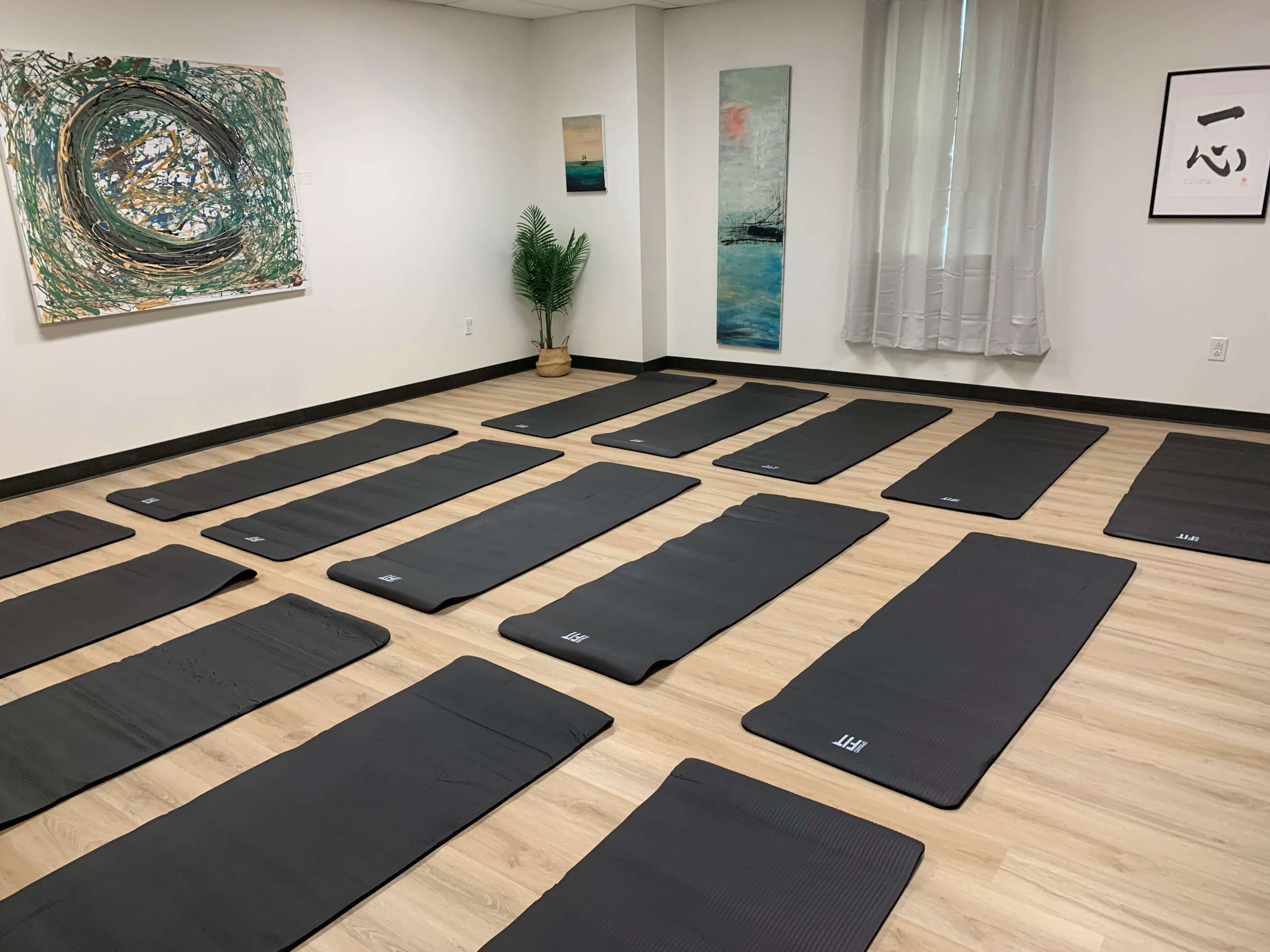 The image shows a yoga studio with a series of black mats neatly arranged on a wooden floor, surrounded by art on the walls and a small plant in the corner.
