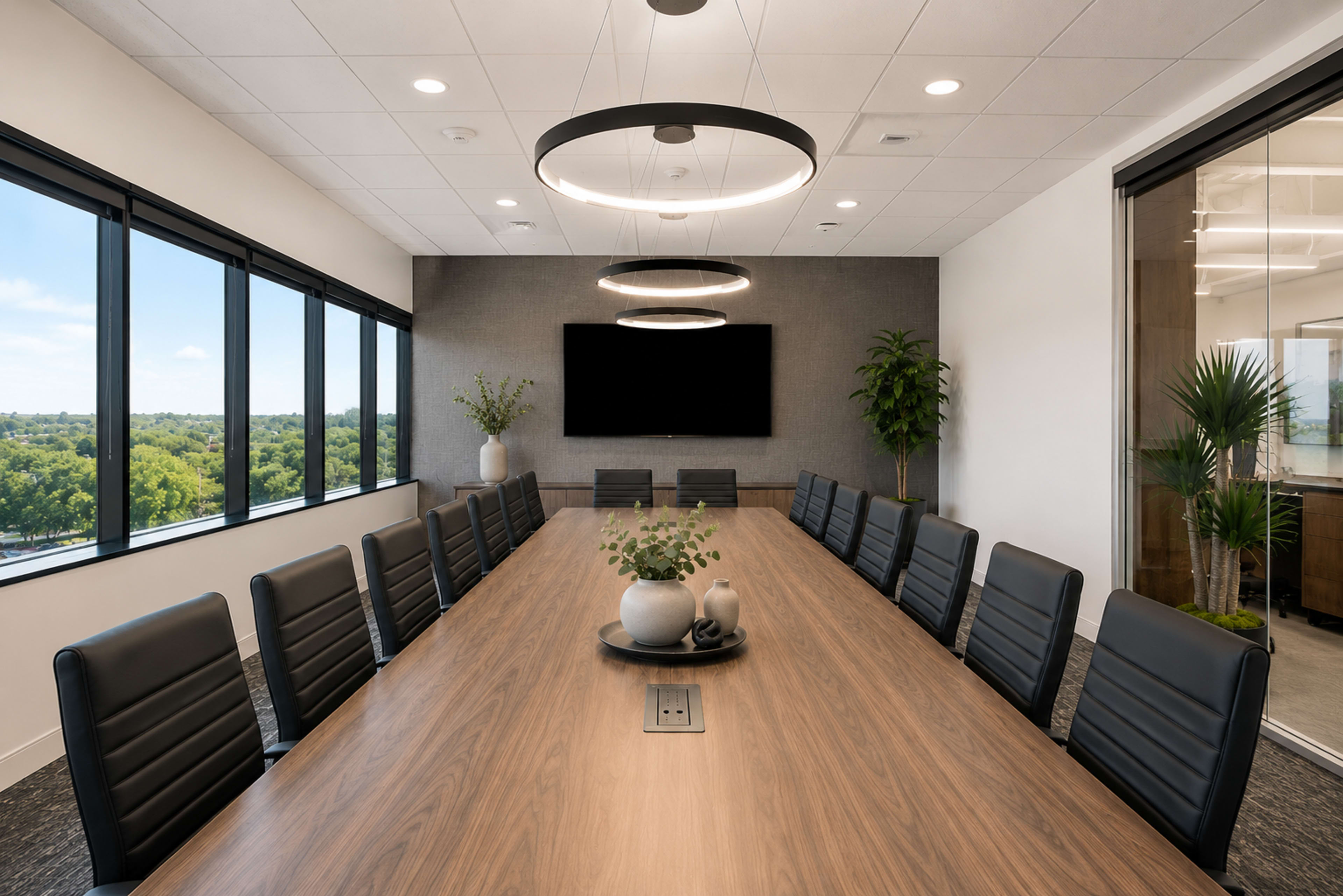 A long wooden conference table is set in a modern meeting room with large windows overlooking a landscape, featuring several black leather chairs and a wall-mounted screen.