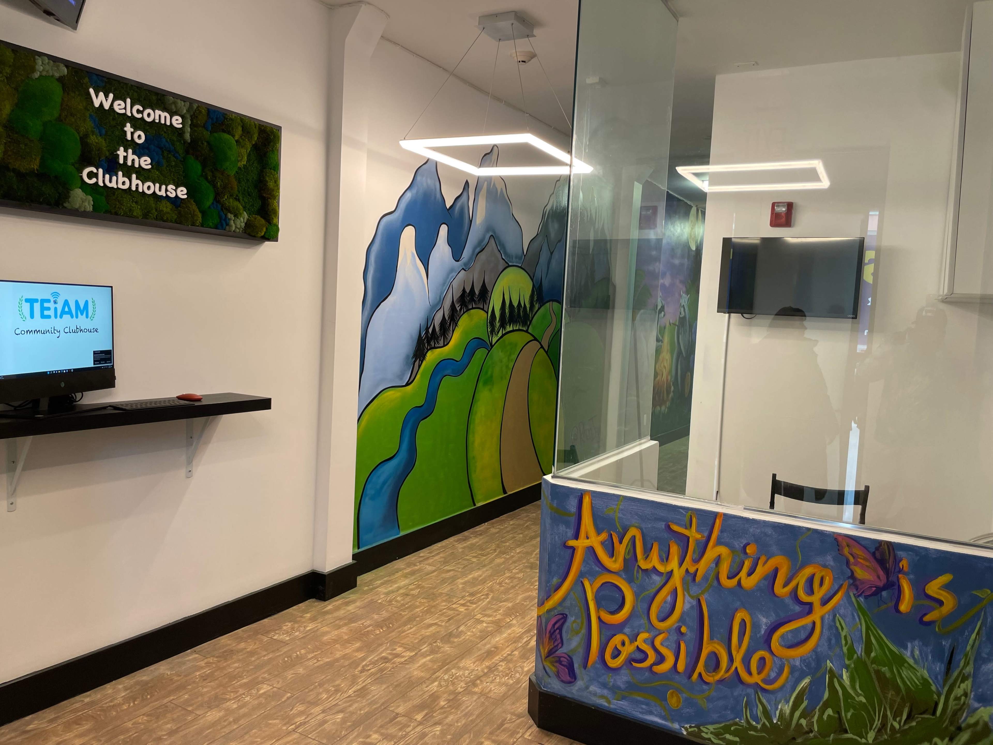 The interior of a community clubhouse features a welcome sign, a colorful mural of mountains and greenery, and a desk with a computer monitor.