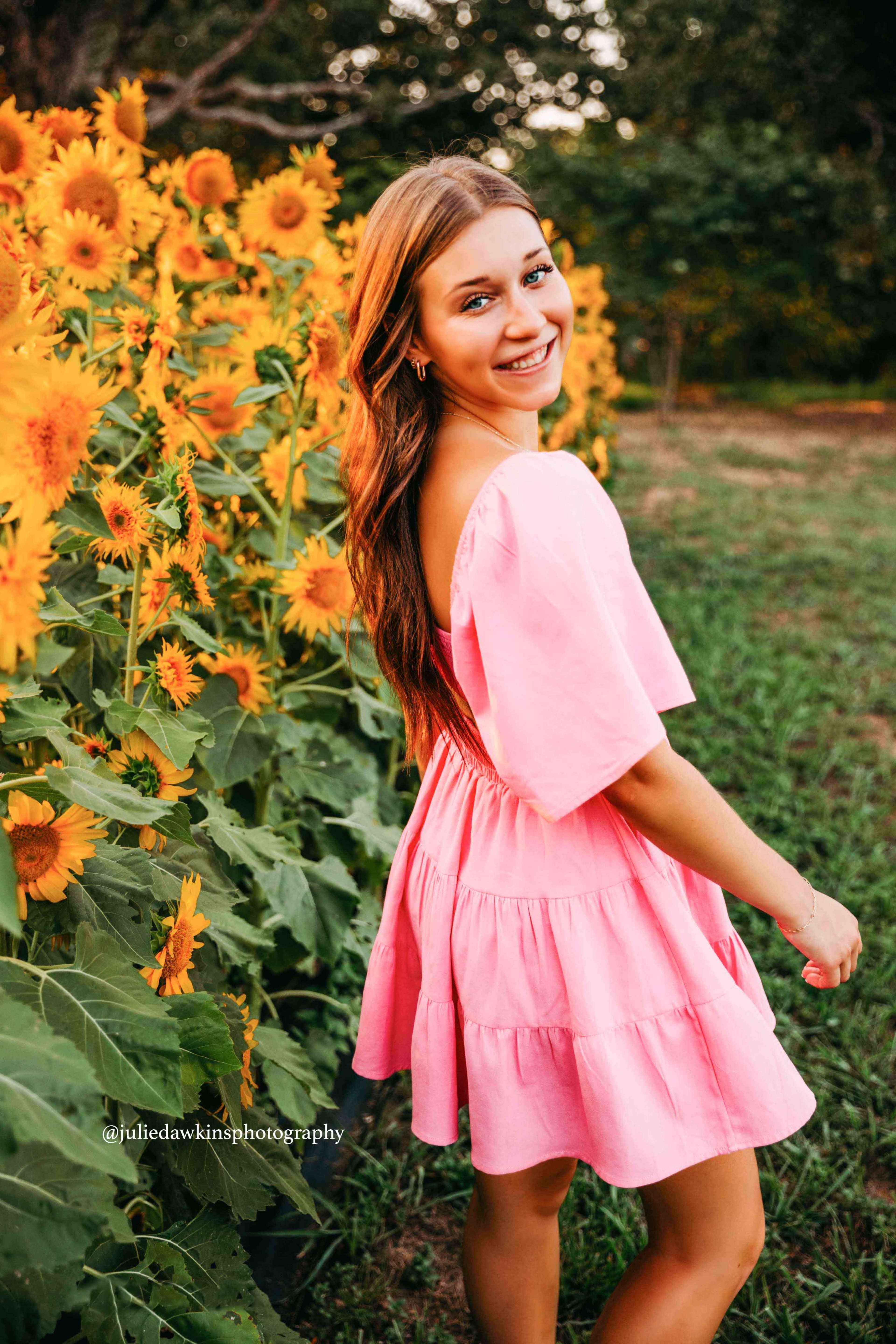 A woman in a pink dress stands among tall sunflowers, smiling as she looks back over her shoulder.