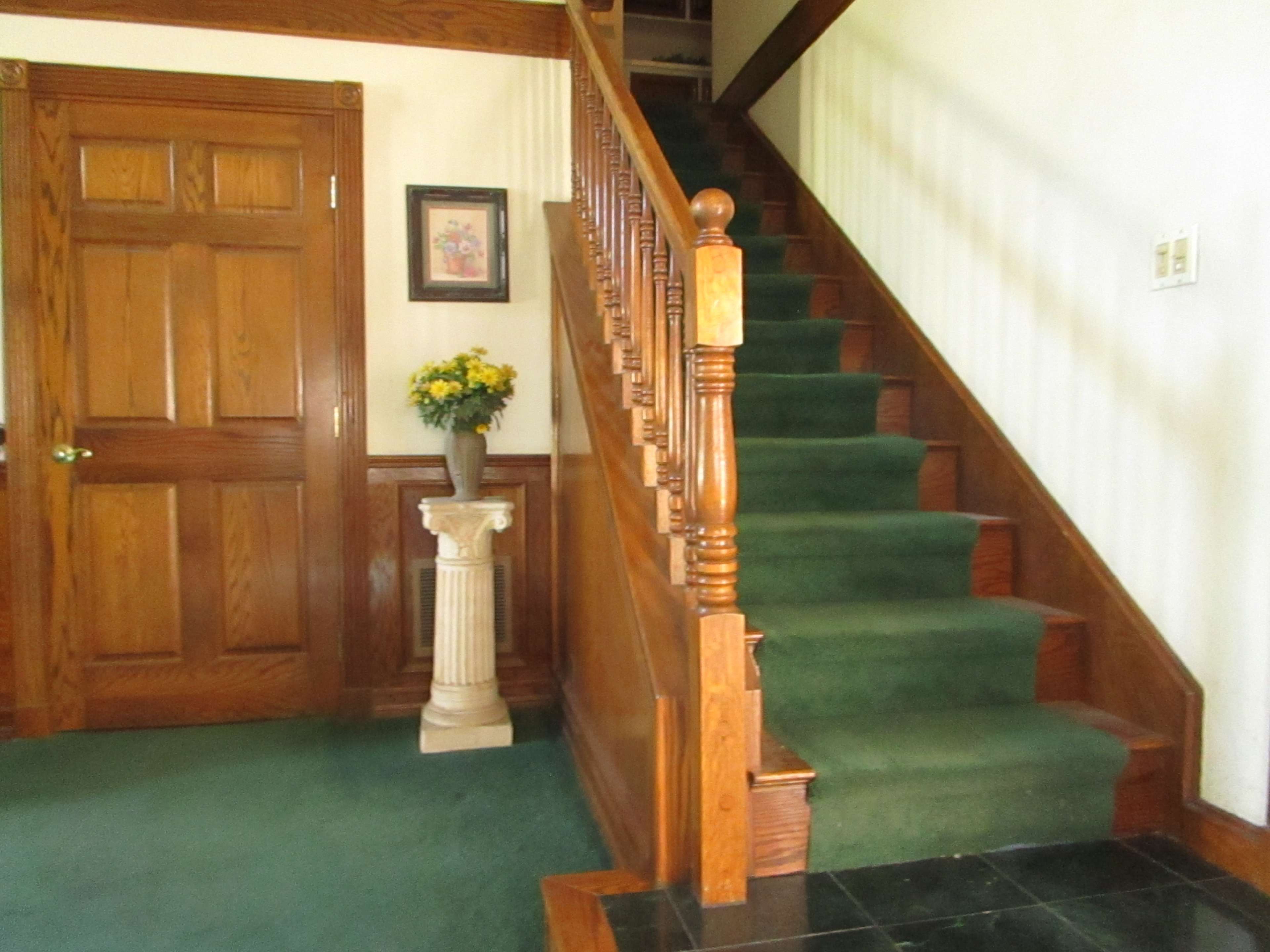 The image features a wooden staircase leading to an upper floor, alongside a door and a decorative column with a flower arrangement.