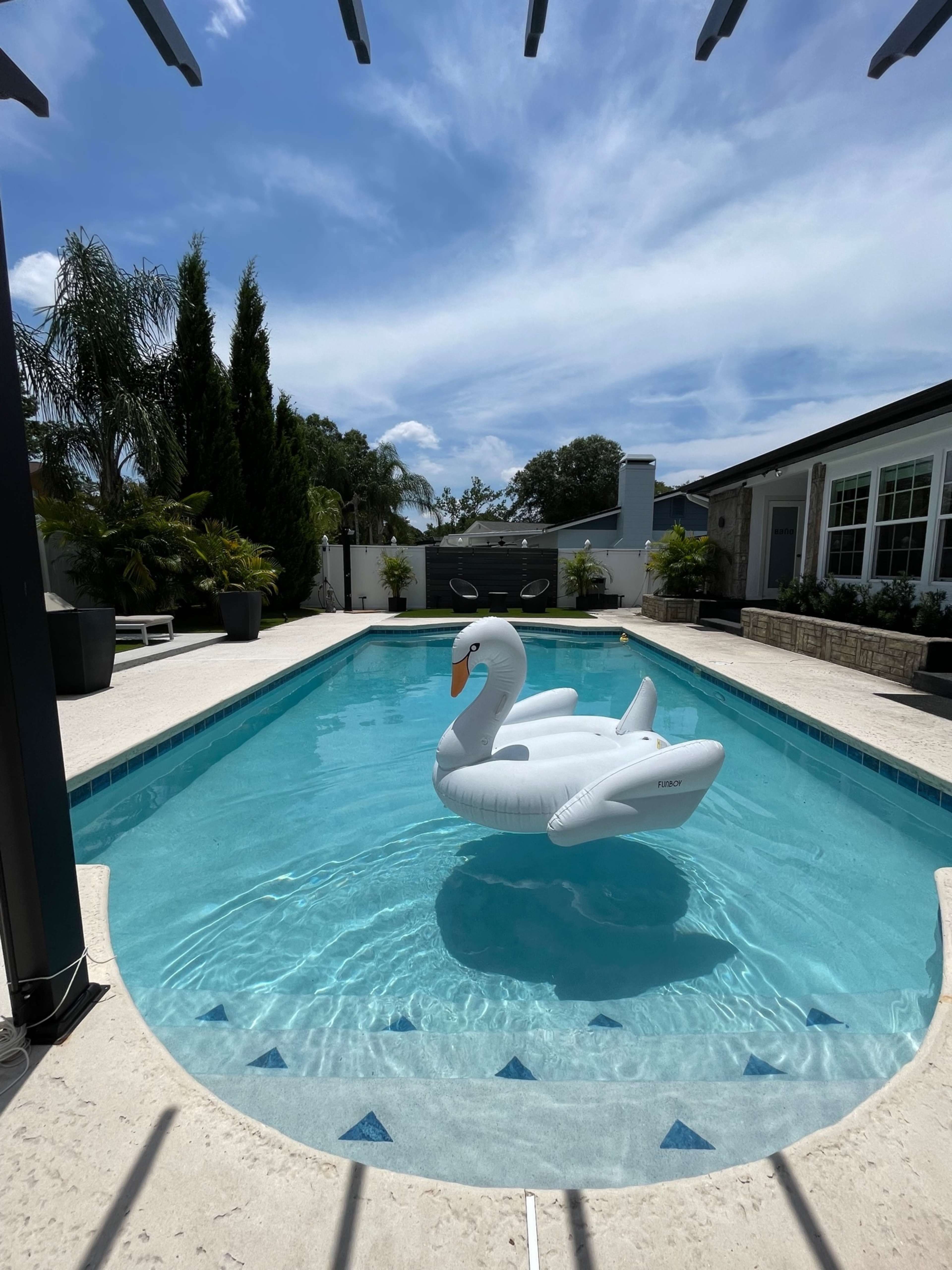 A large inflatable swan floats on a clear blue swimming pool surrounded by palm trees and a modern backyard.