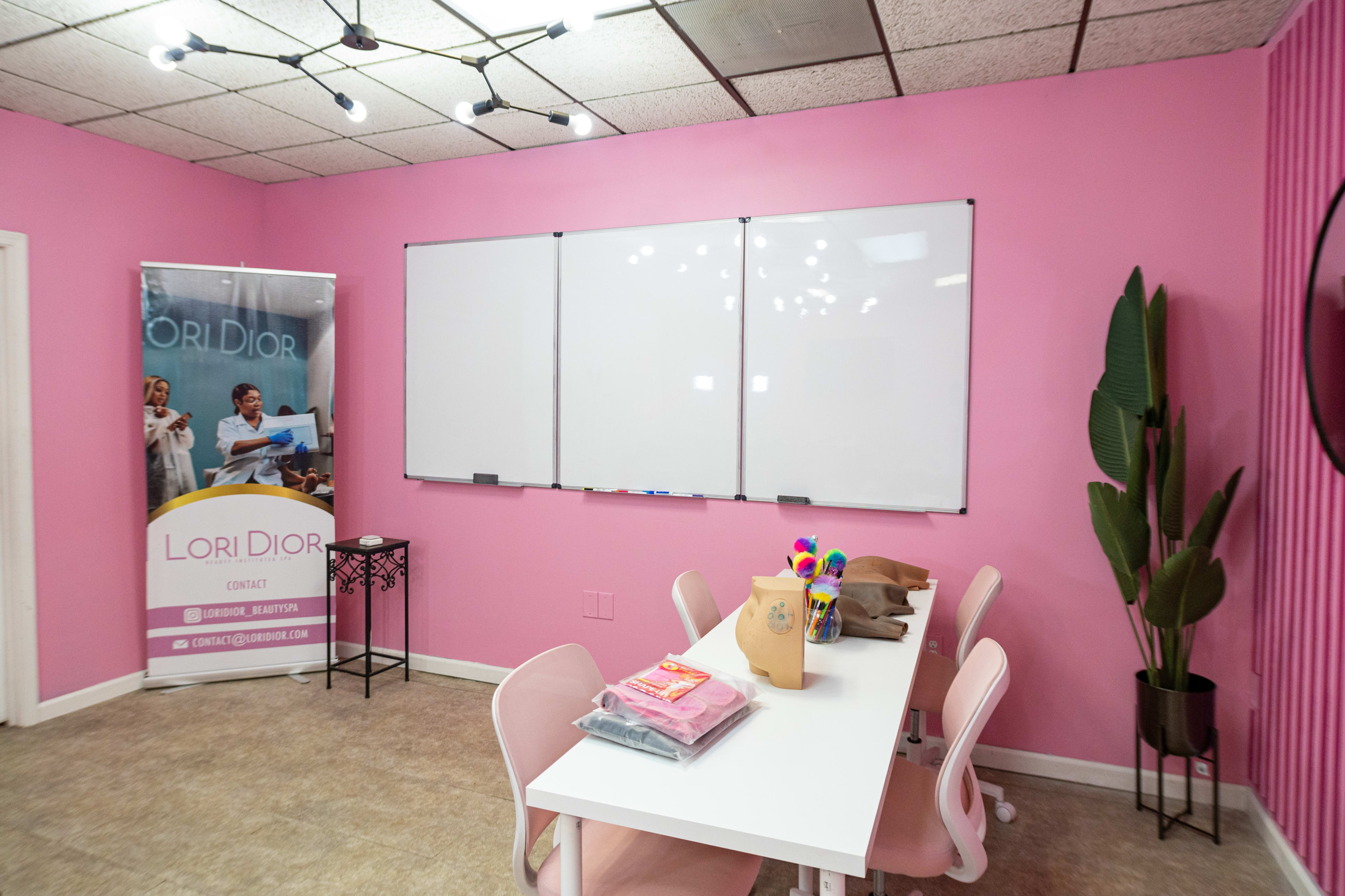 A pink-walled meeting room features a white table with chairs, a large whiteboard, and a promotional banner for Lori Dior.