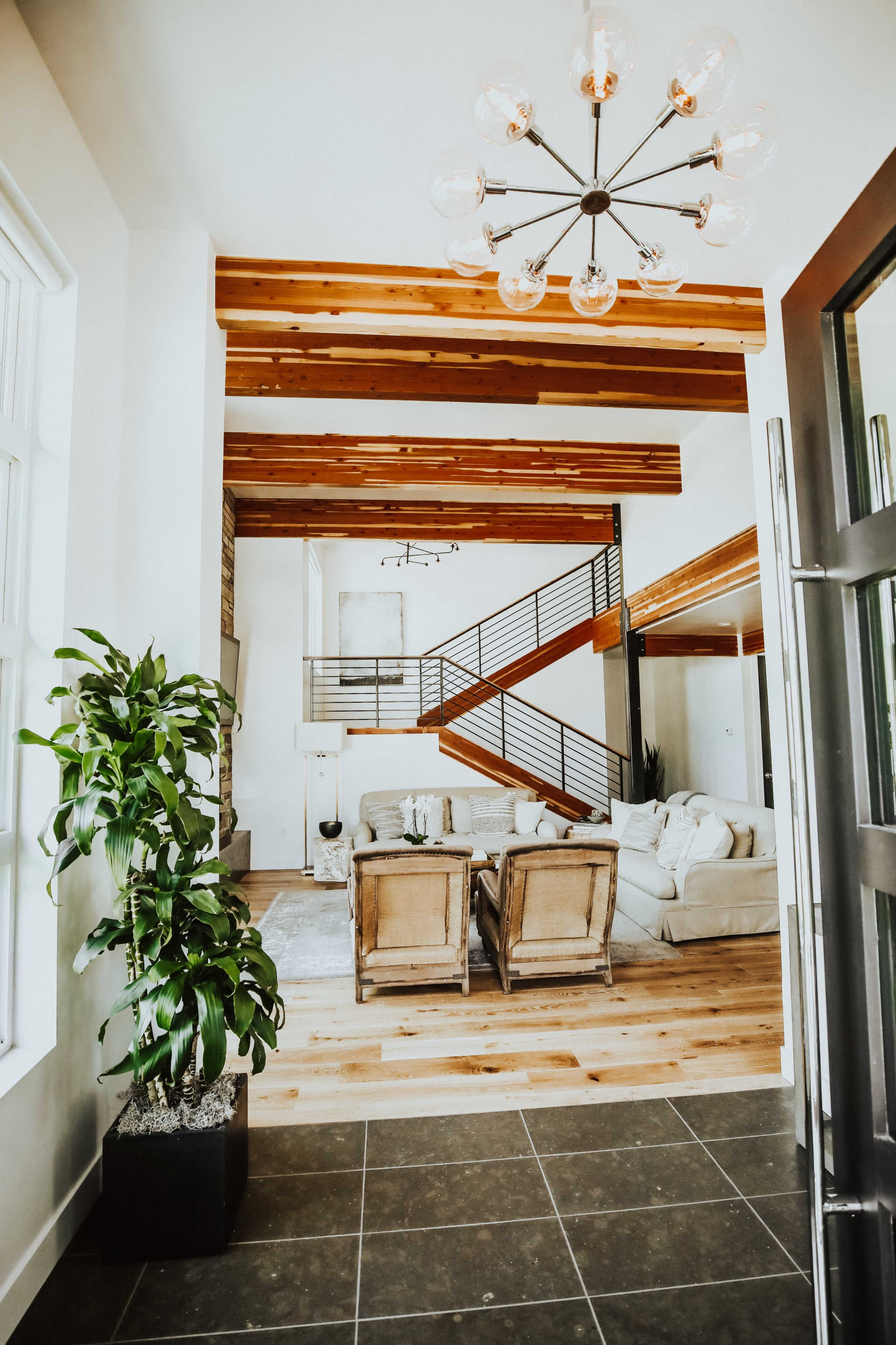 A welcoming entryway that opens into a spacious living area with a staircase, a light fixture, and a cozy seating arrangement surrounded by wooden beams and large windows.