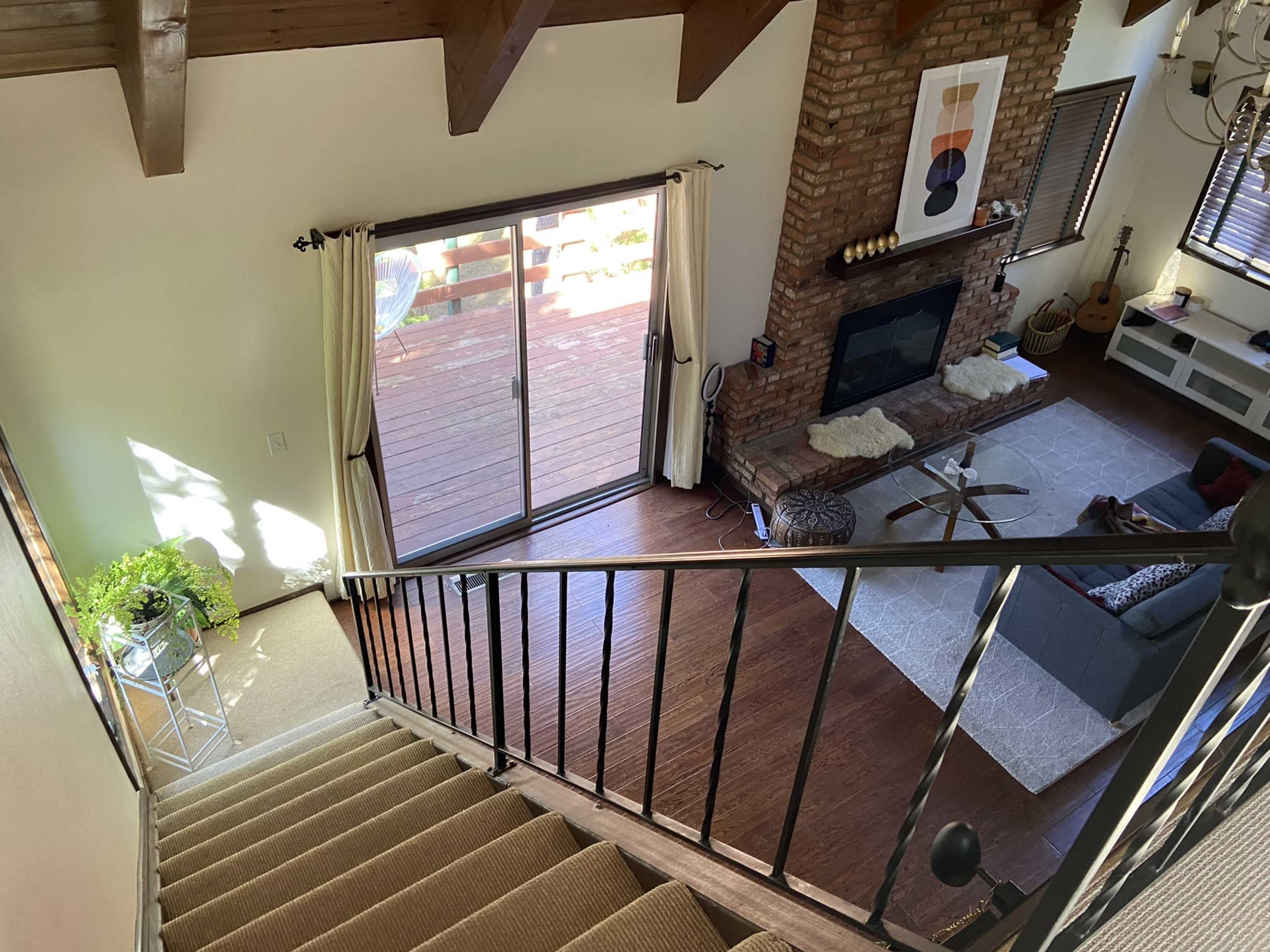 A staircase descends into a living room with a view of a deck through sliding glass doors, featuring a brick fireplace and a large area rug.