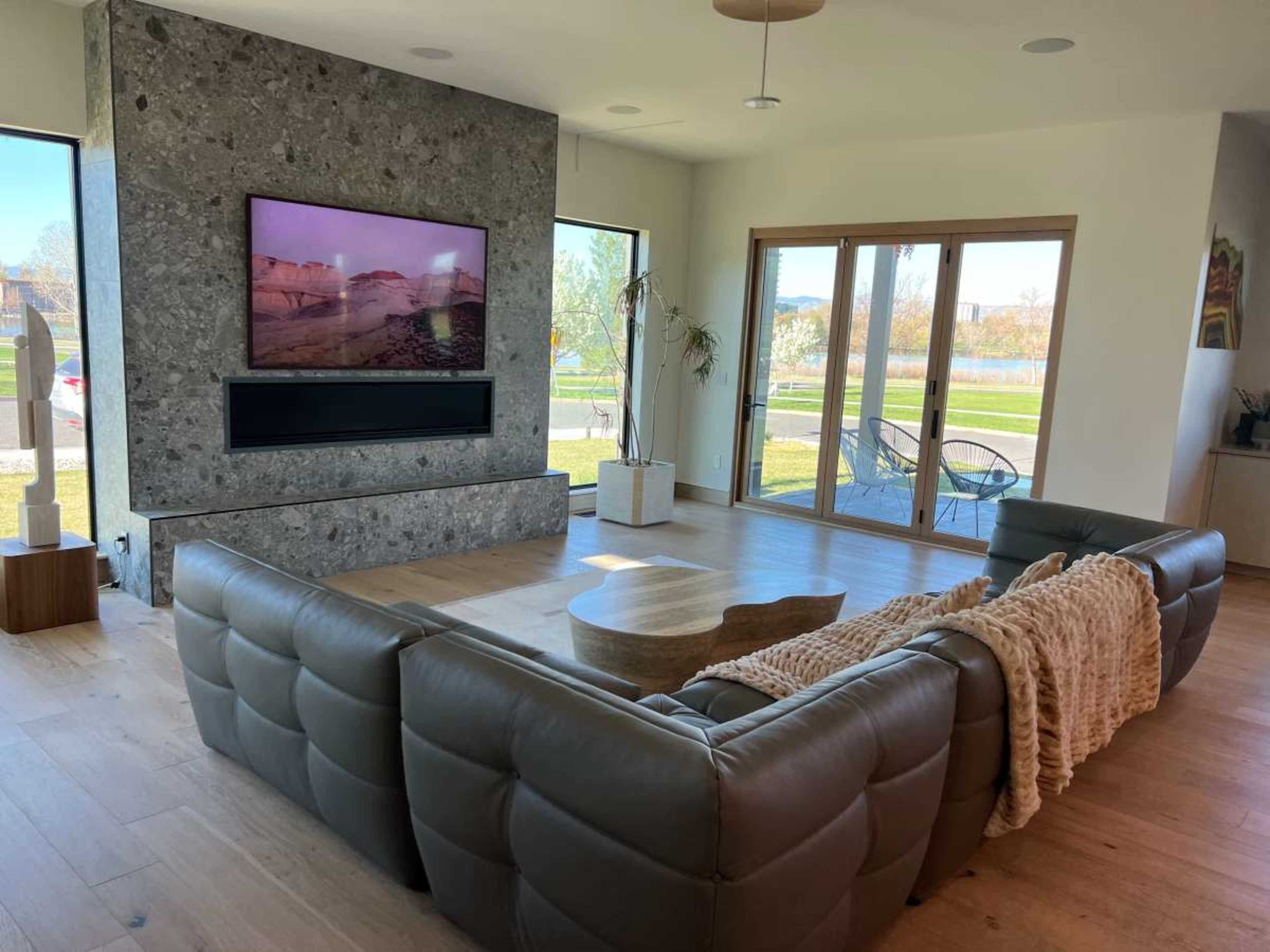 A modern living room features a large sectional couch facing a wall-mounted television, with large windows offering a view of an outdoor landscape.