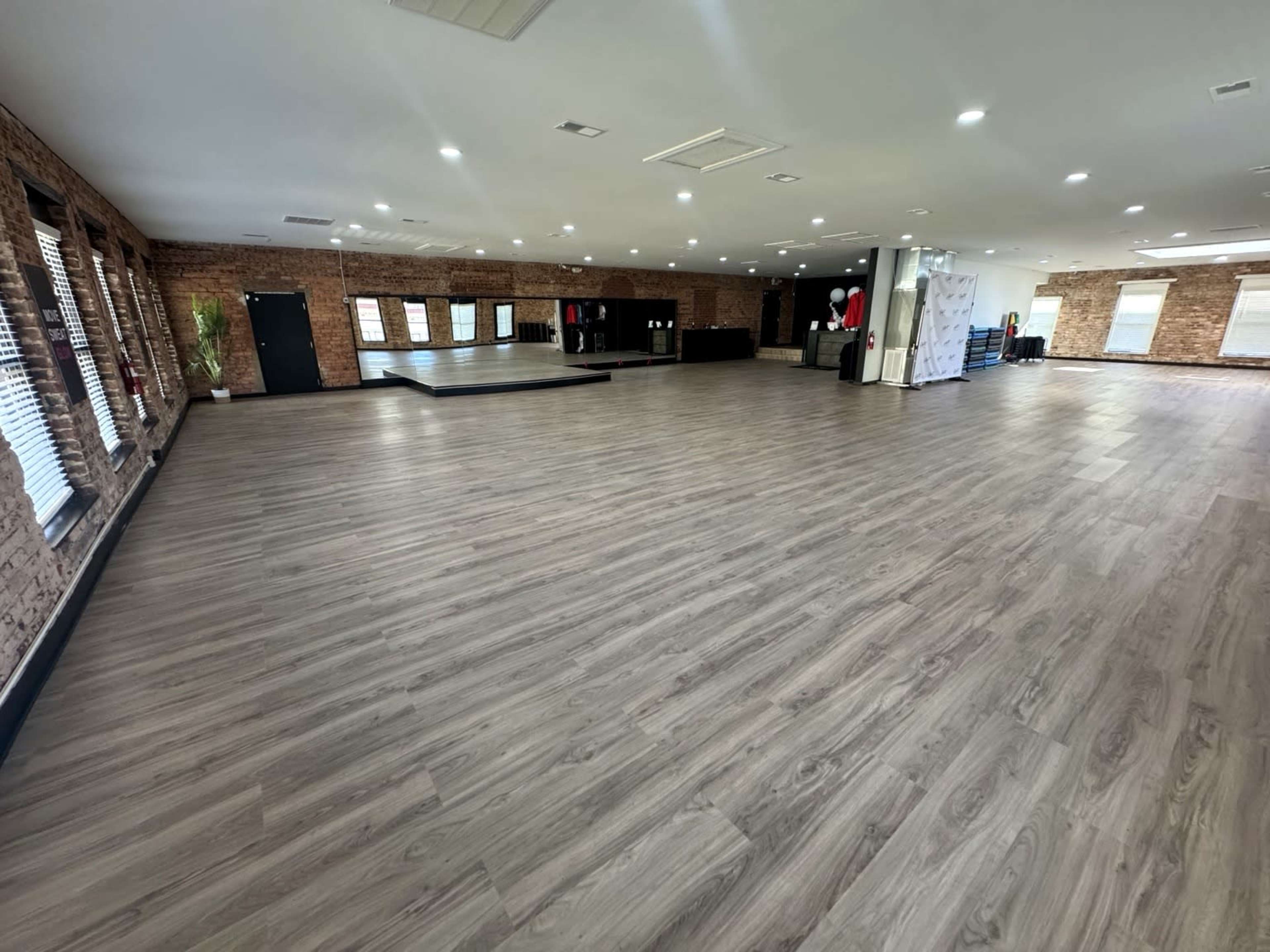 The image shows a spacious interior room with wooden flooring, large windows, and exposed brick walls, featuring a stage area in the background.