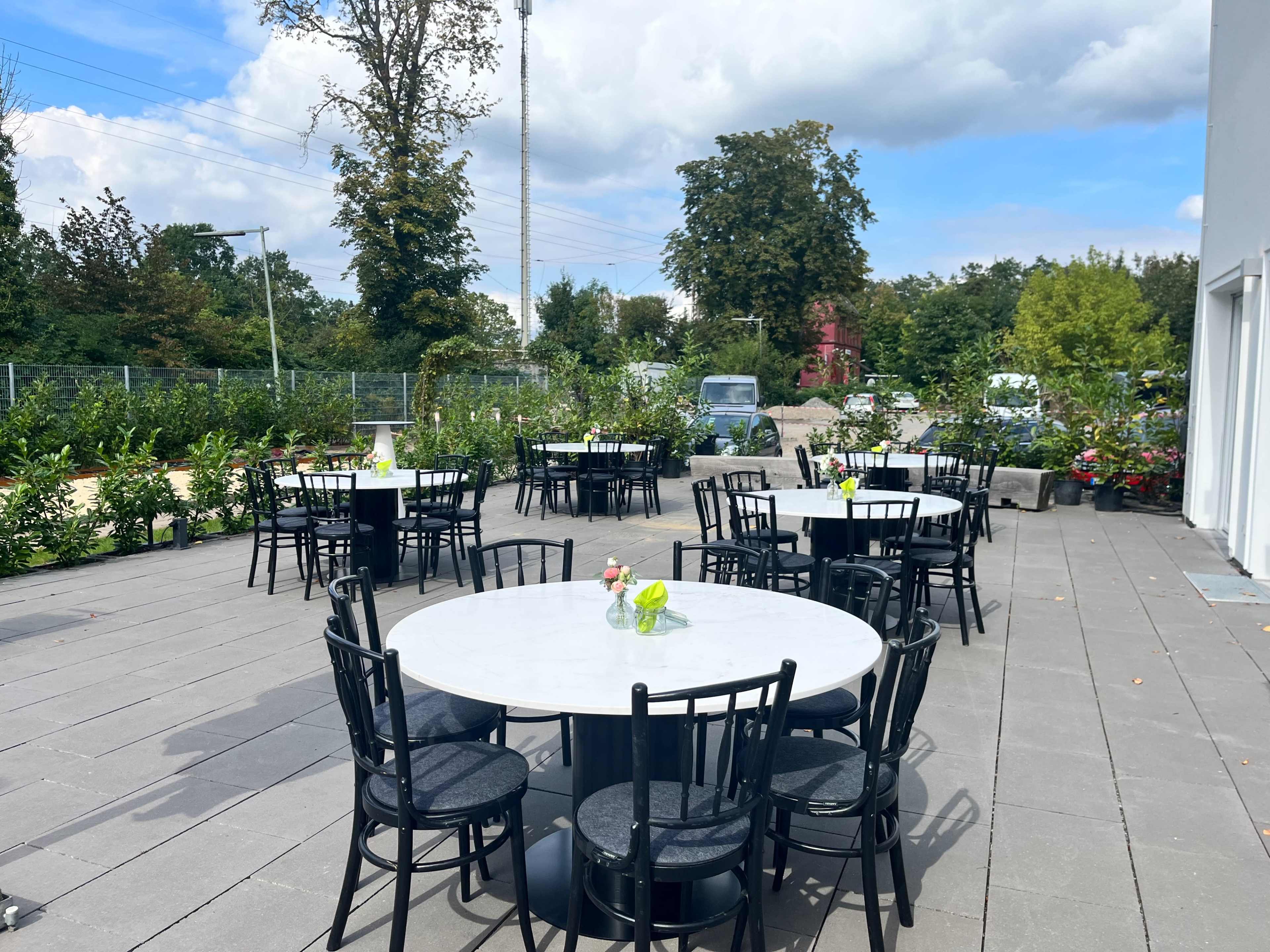 A spacious outdoor patio features several round tables with marble tops and black chairs, surrounded by greenery and under a partly cloudy sky.