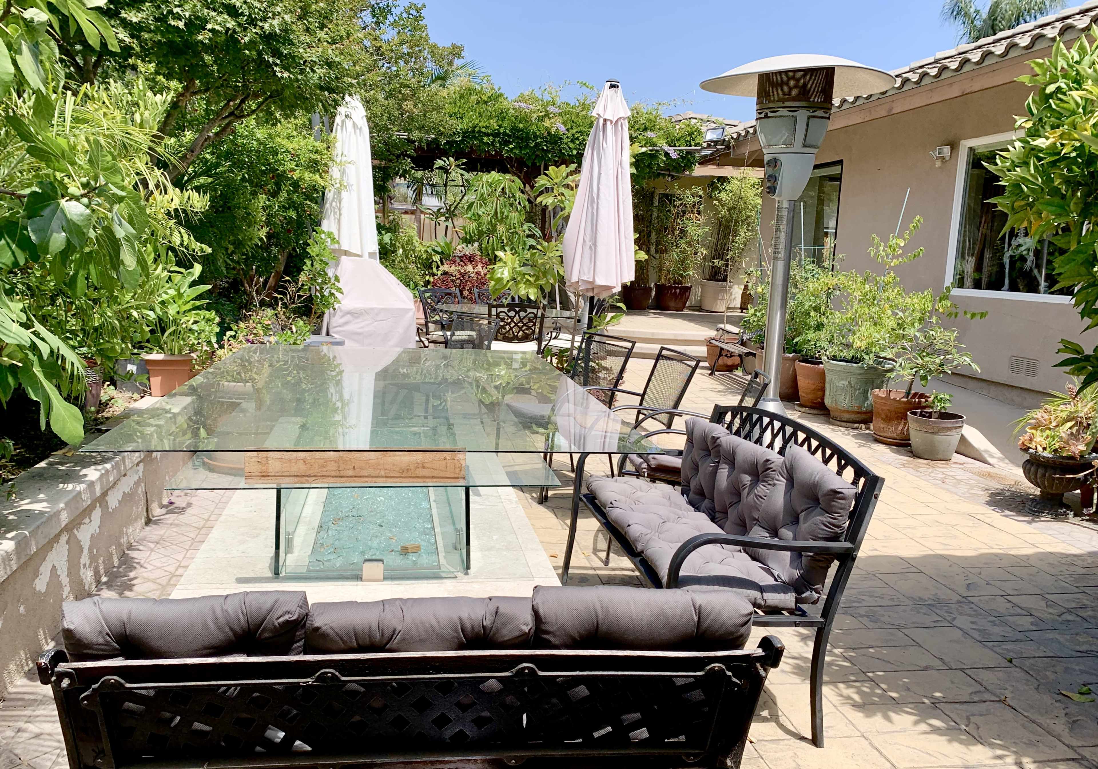 A patio area features a glass-top table surrounded by black metal chairs and sofas, with greenery and potted plants in the background.