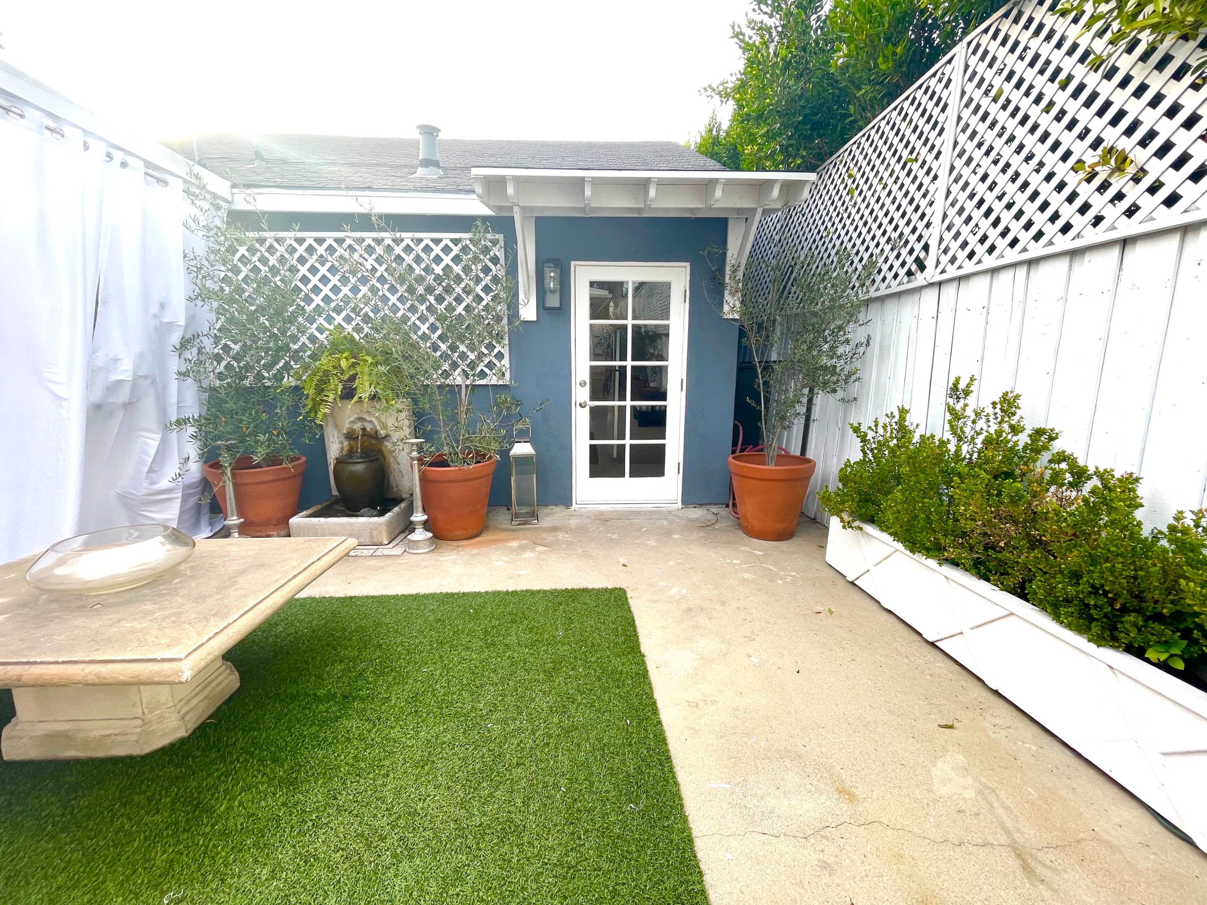 The image shows a small outdoor patio area featuring a green artificial grass surface, potted plants, and a door leading to a building.