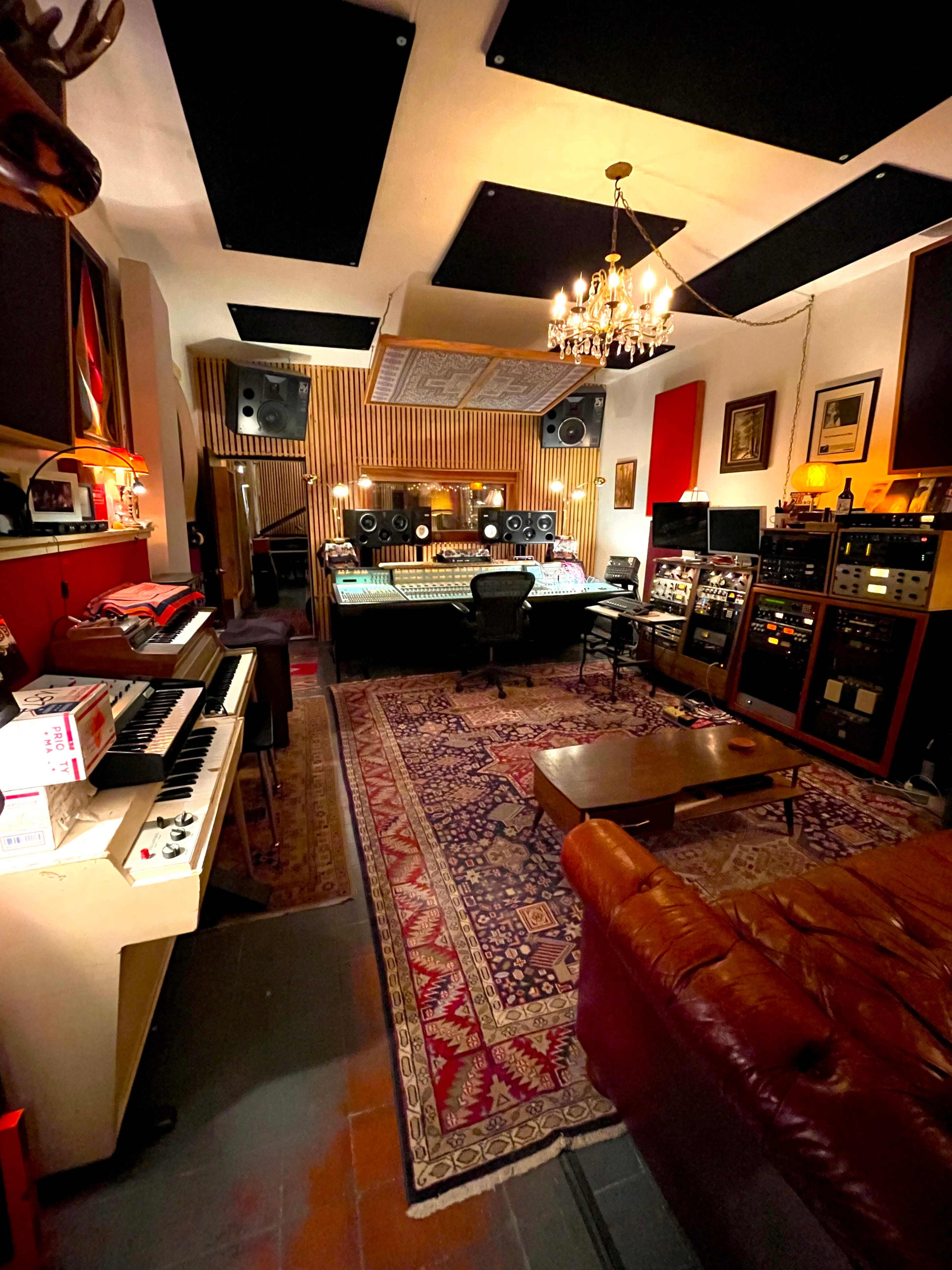 A recording studio with a large mixing console, various audio equipment, a keyboard, and a plush sofa on a patterned rug.
