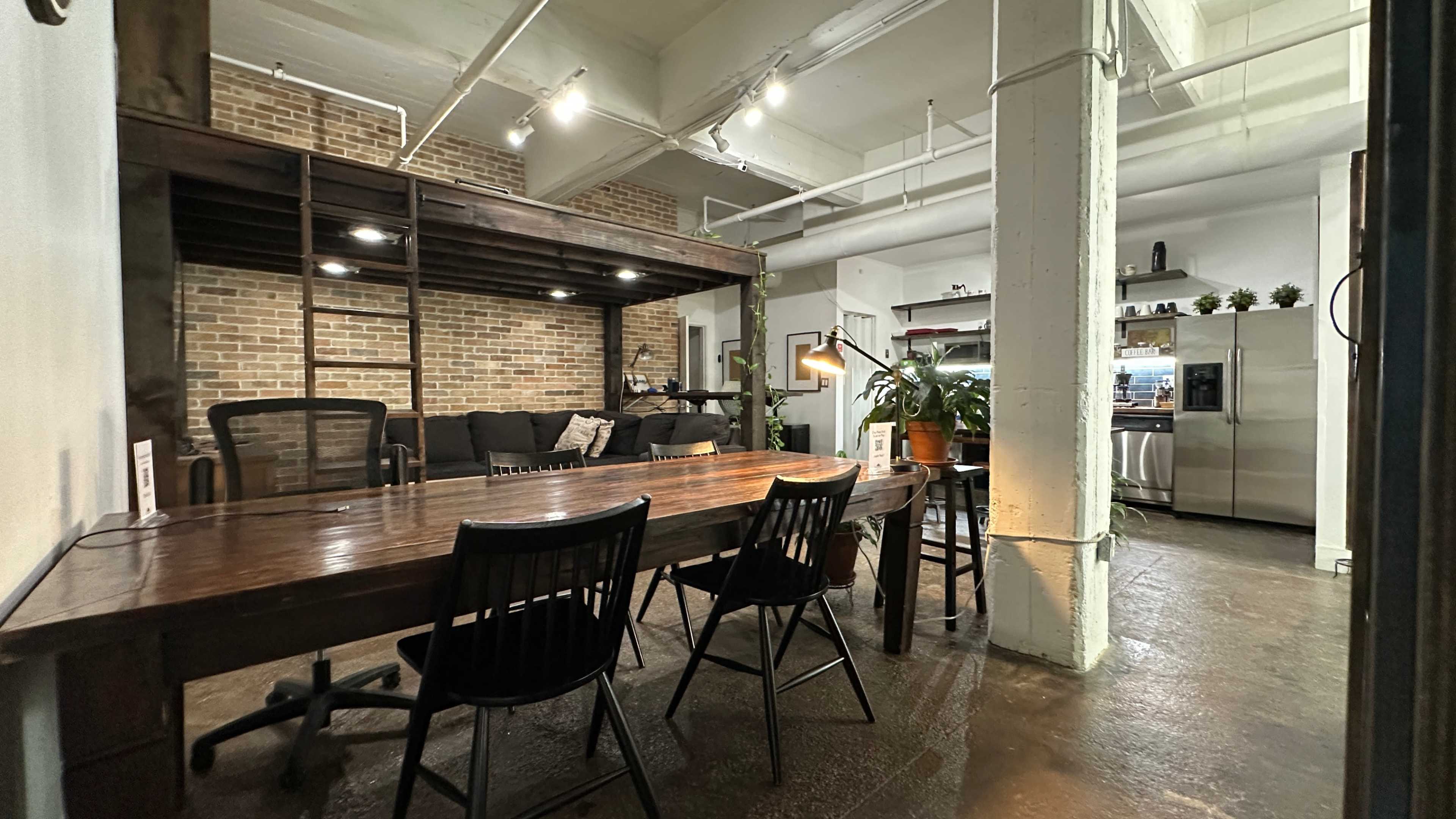 The image shows an industrial-style office space with a large wooden table, several black chairs, exposed brick walls, and a kitchen area in the background.