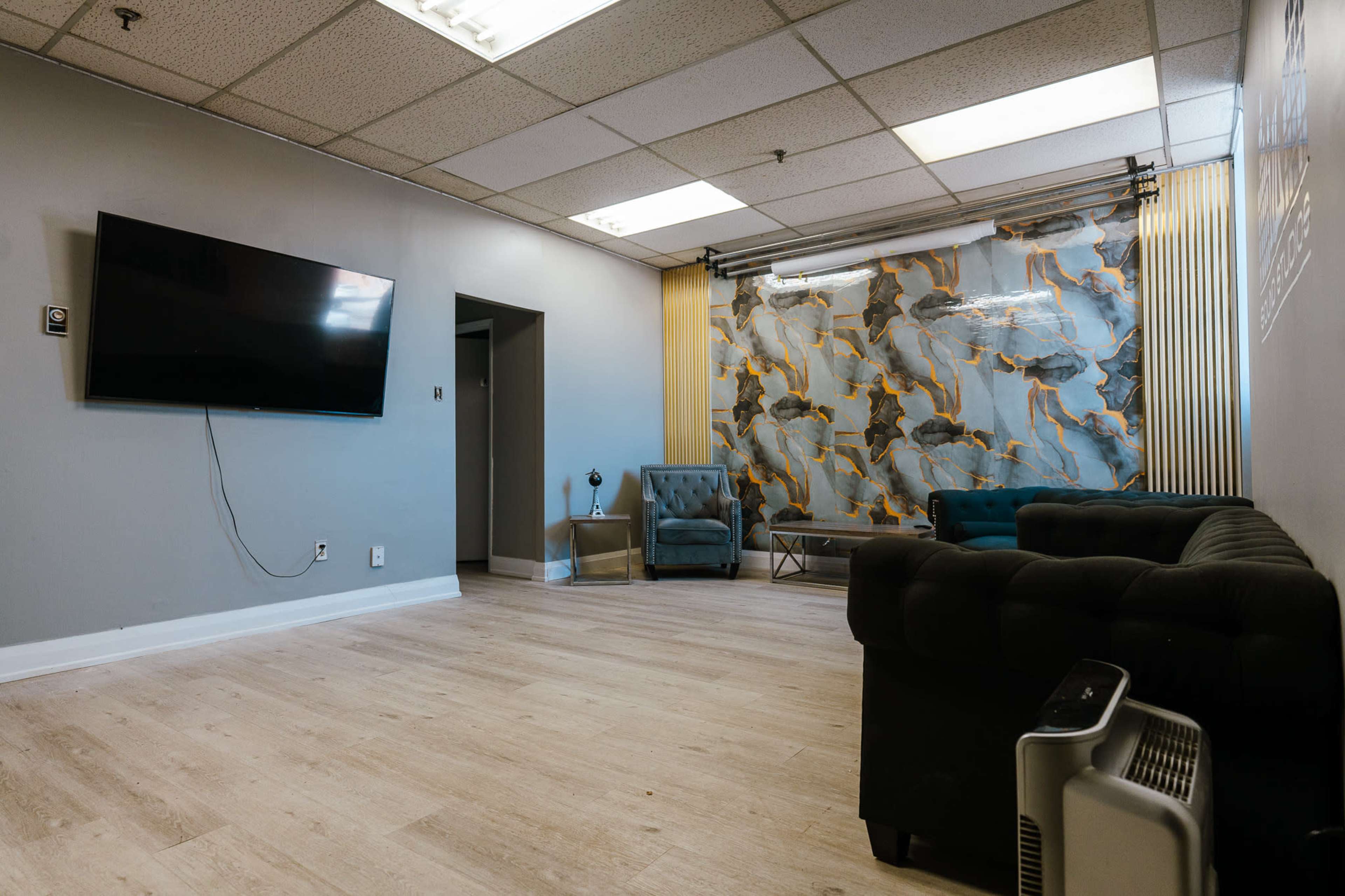 The image shows a spacious, minimalist room with a large television mounted on the wall, a patterned backdrop, and seating arrangements including a sofa and armchairs.