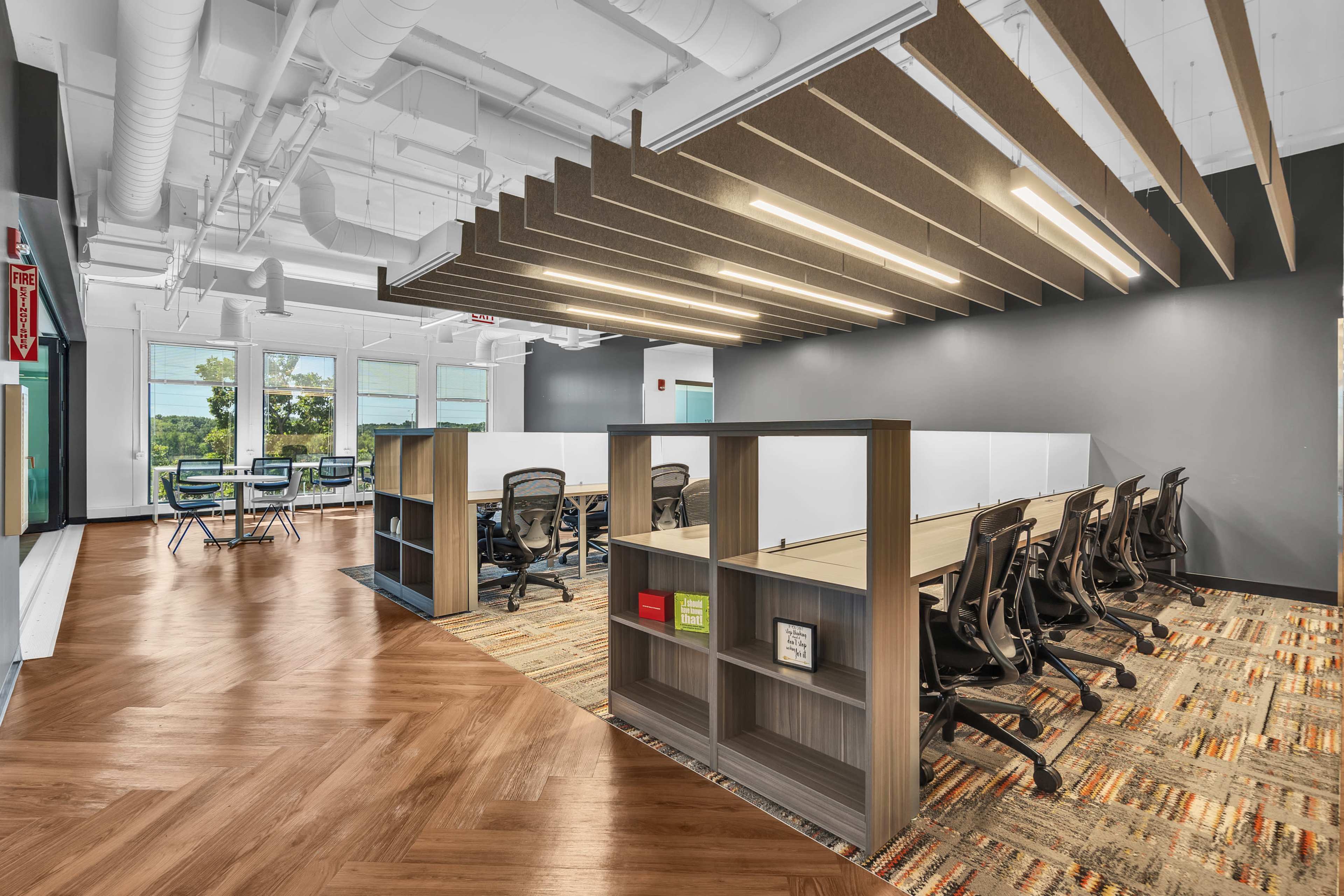 The image shows a modern office space featuring rows of desks with ergonomic chairs, wooden partitioning, and a patterned area rug under overhead lighting.