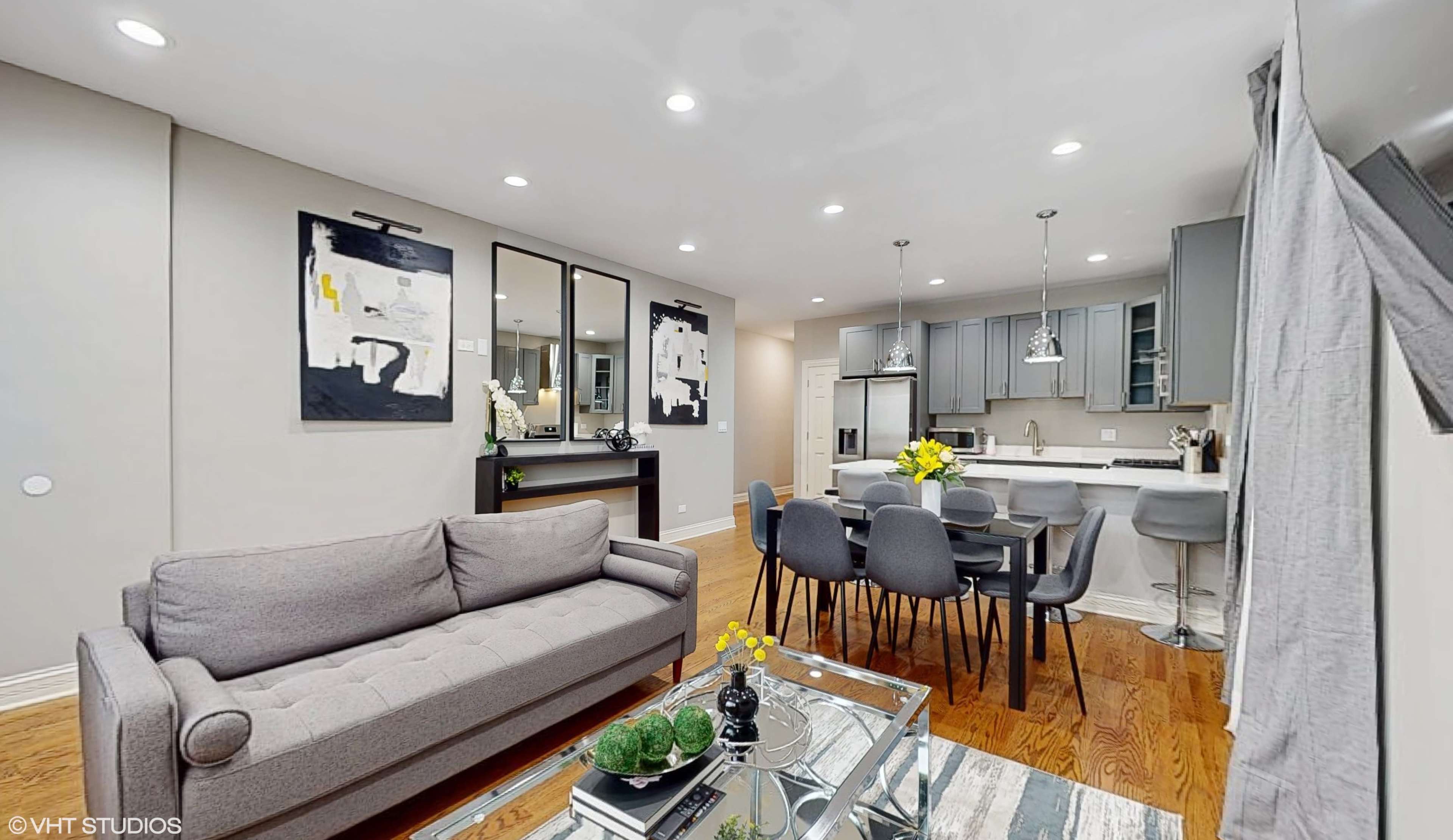 A modern living and dining area with a gray sofa, a dining table surrounded by gray chairs, and a kitchen area in the background.