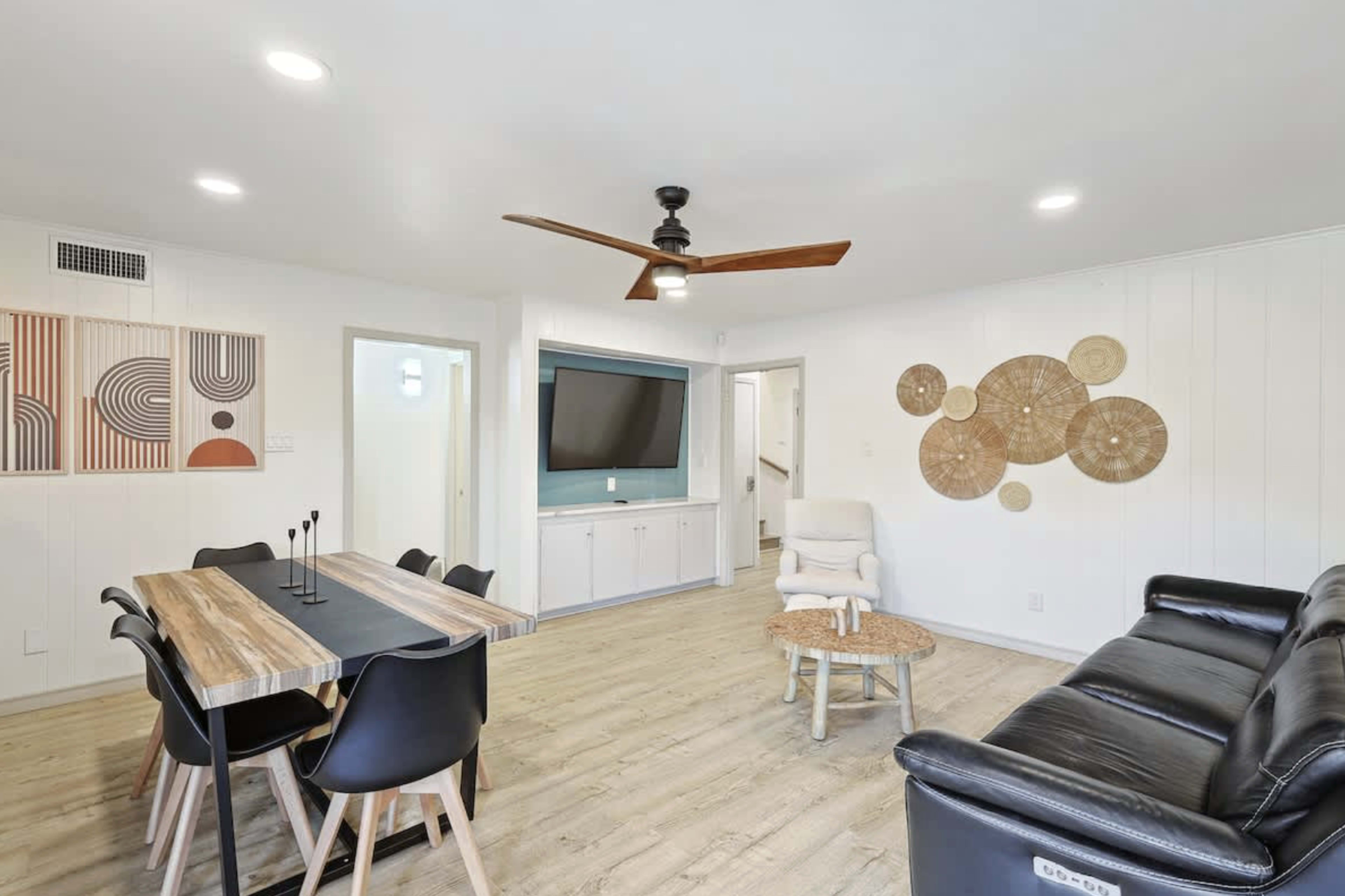 The image shows a living and dining area featuring a wooden table with black chairs, a leather sofa, a wall-mounted television, and decorative wall art.