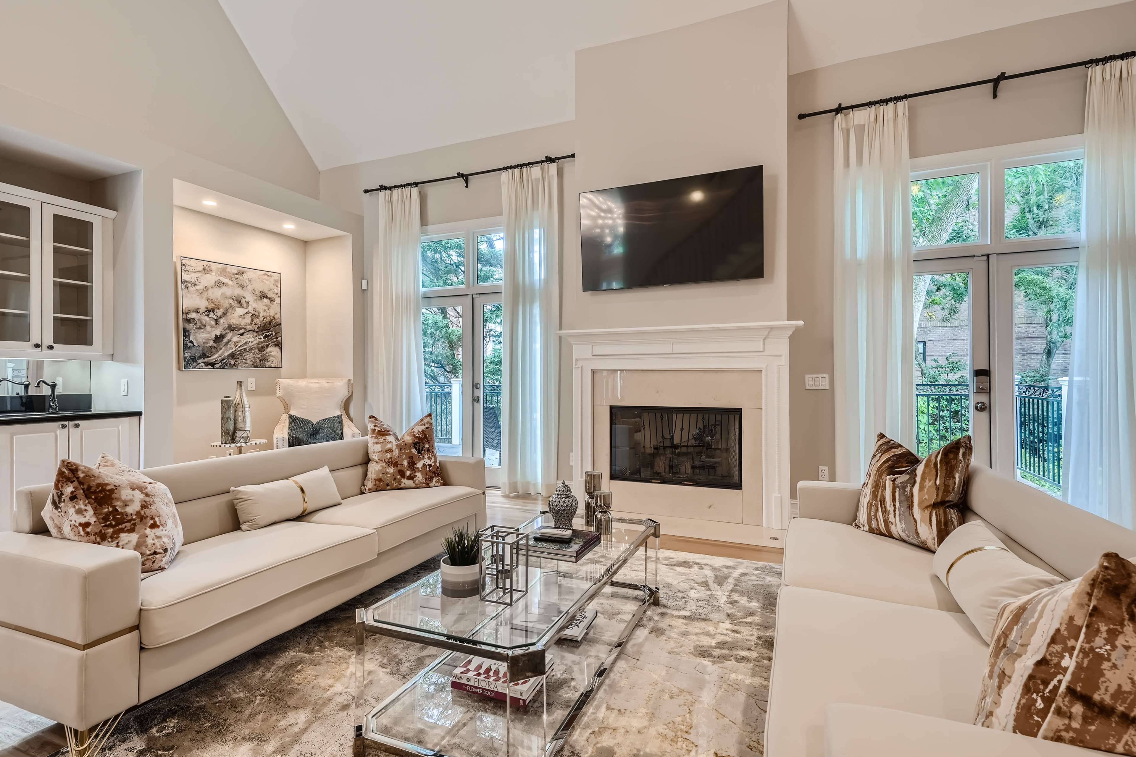 The living room features two white sofas with decorative pillows, a glass coffee table, and large windows overlooking a garden.