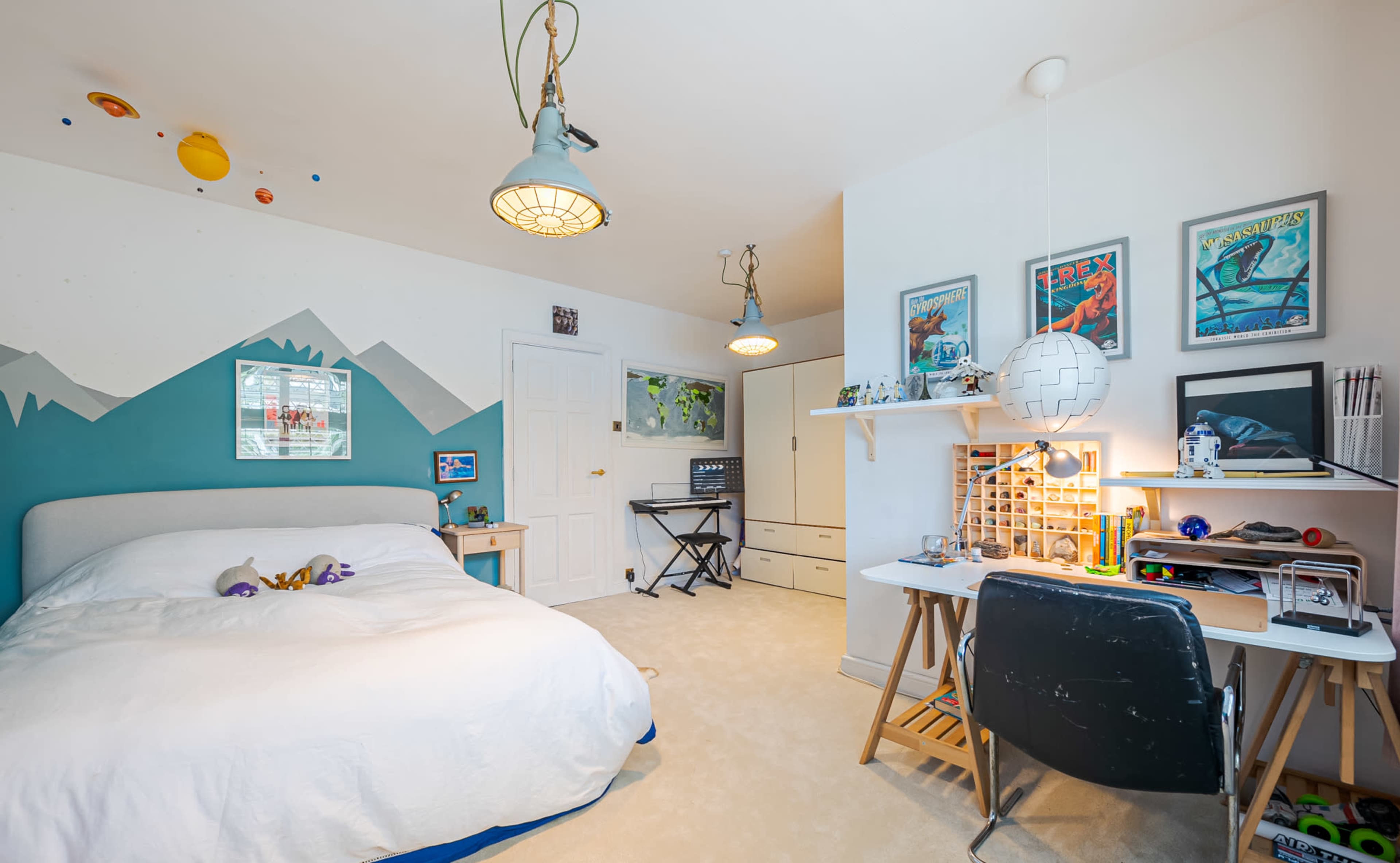 A bedroom with a bed, a desk with a lamp and art supplies, and a wall decorated with mountain graphics and framed posters.