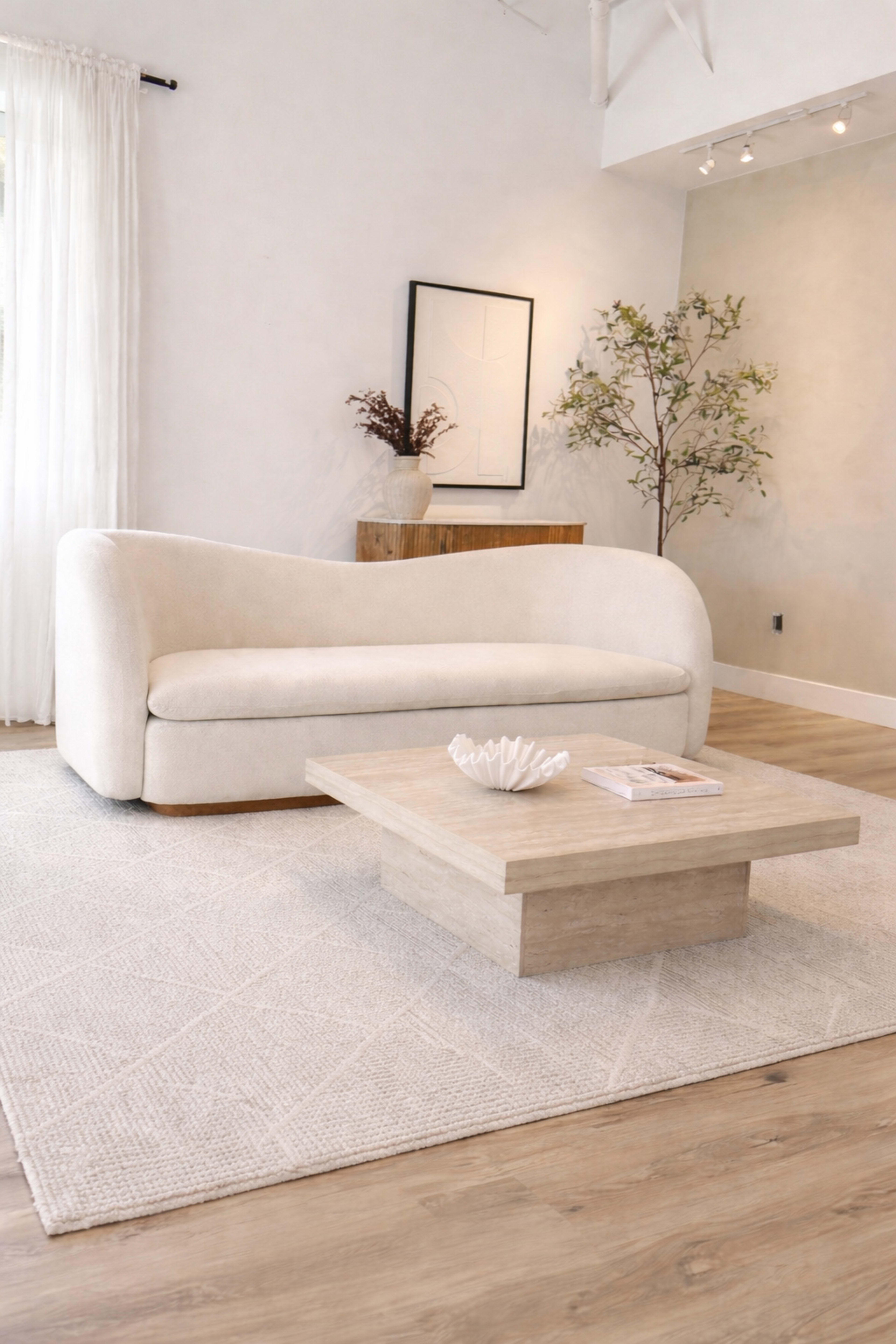 The image shows a light-colored, modern living room featuring a curved sofa, a simple wooden coffee table, a decorative bowl on the table, and greenery in a corner.