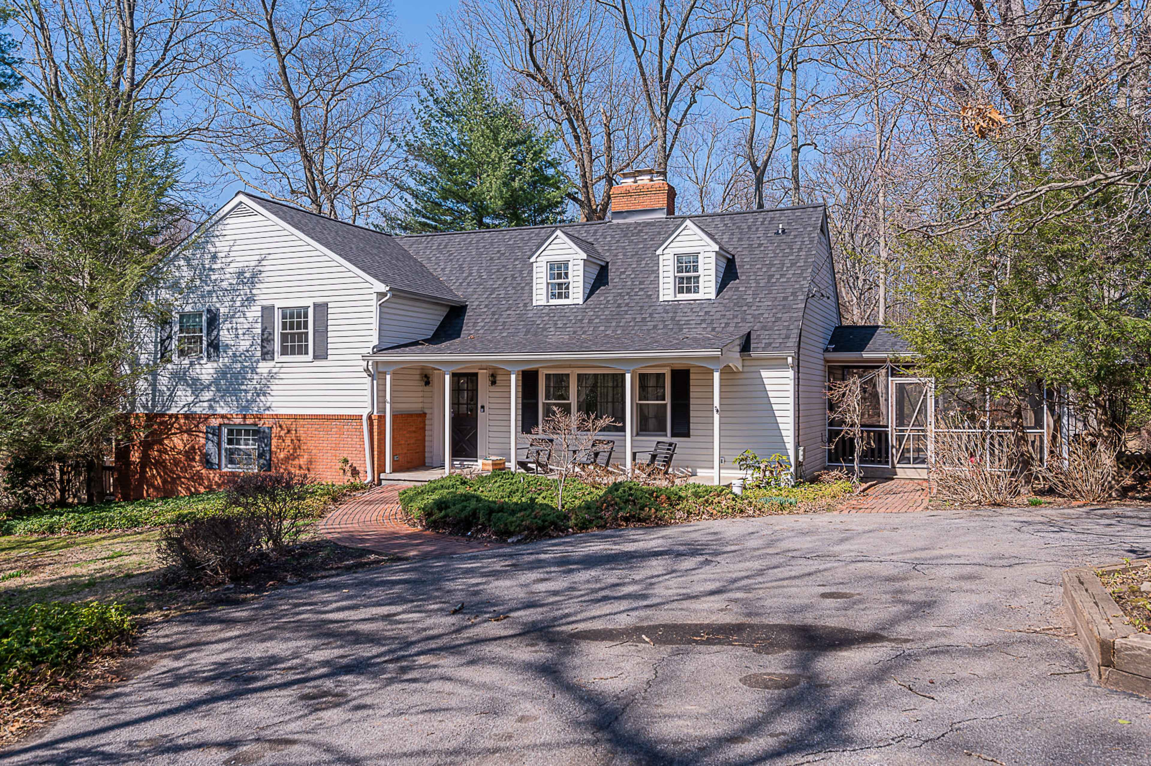 The image shows a two-story white house with a black roof and a brick front porch, surrounded by trees and a gravel driveway.