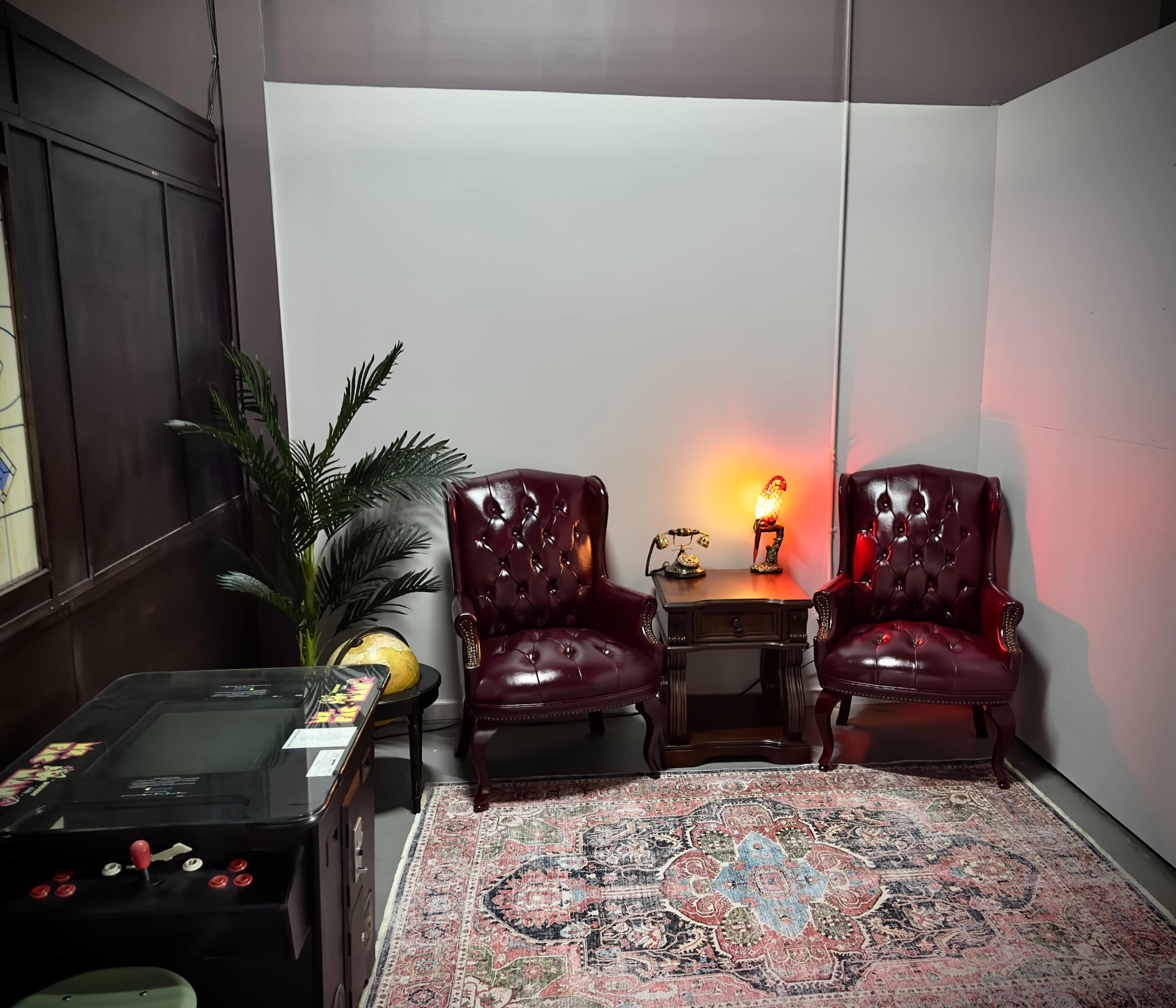A small room featuring two burgundy armchairs, a wooden table with a lamp, a decorative plant, and a vintage arcade game along with a patterned rug.
